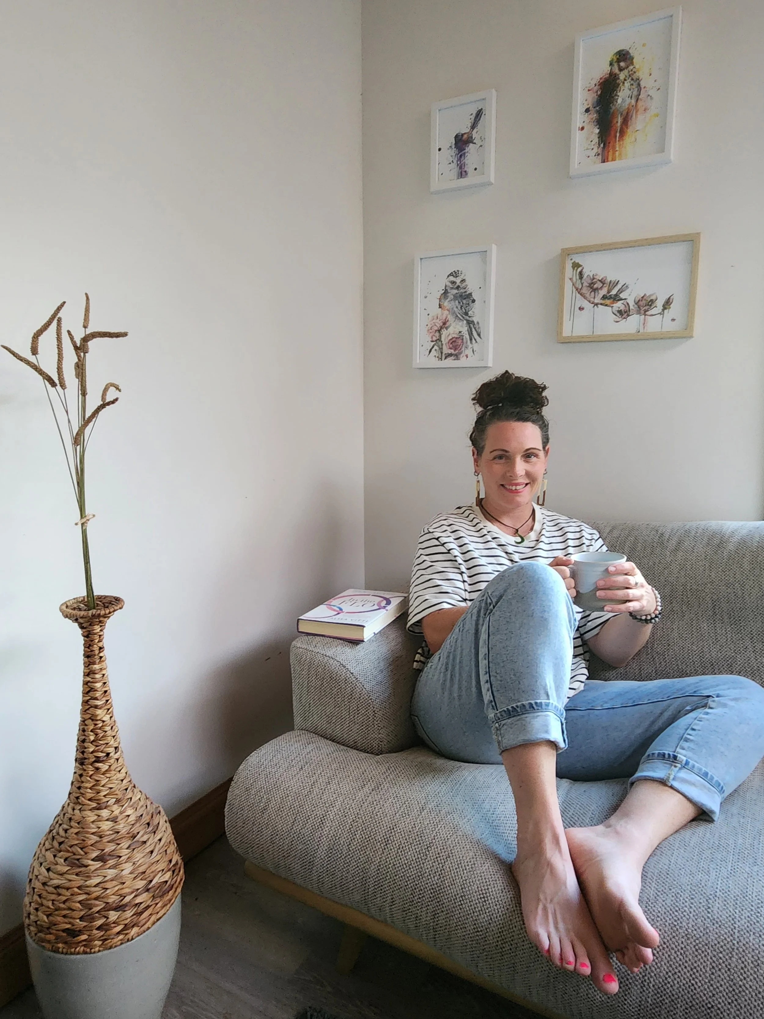 A woman sitting on a beige sofa with a cup in her hand in a living room, with artwork on the wall behind her and a tall wicker vase with dried plants beside her.