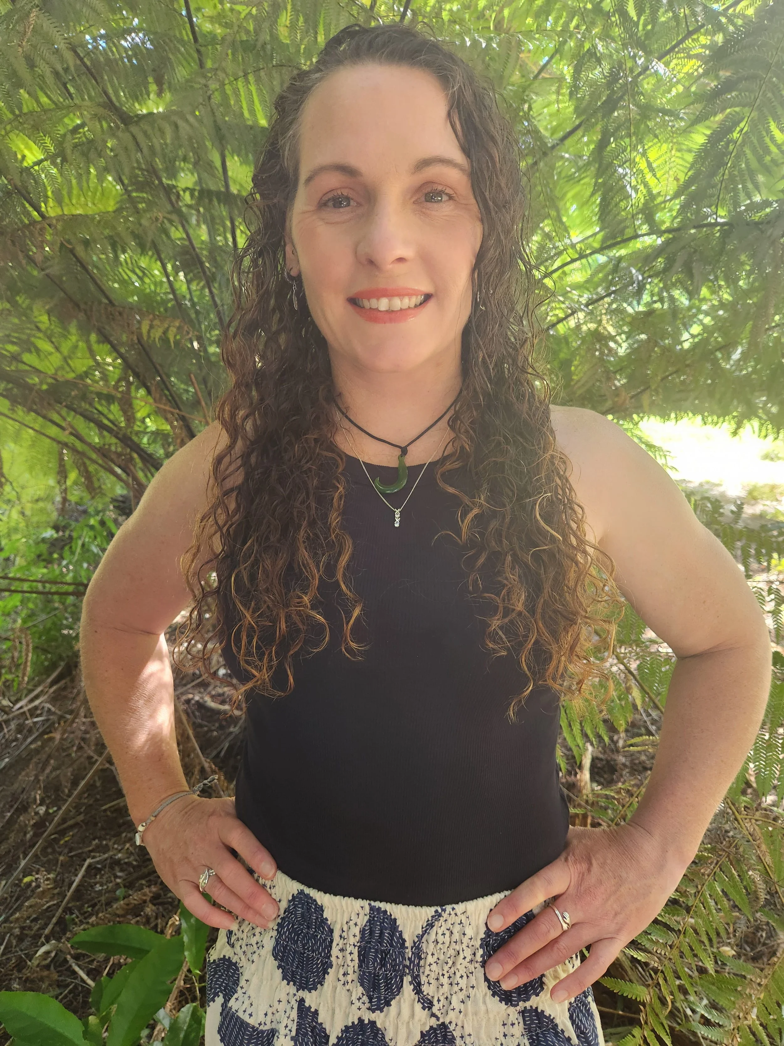 A woman with long, curly brown hair, smiling, standing outdoors in front of greenery. She wears a black sleeveless top, patterned shorts, and jewelry including necklaces and rings.