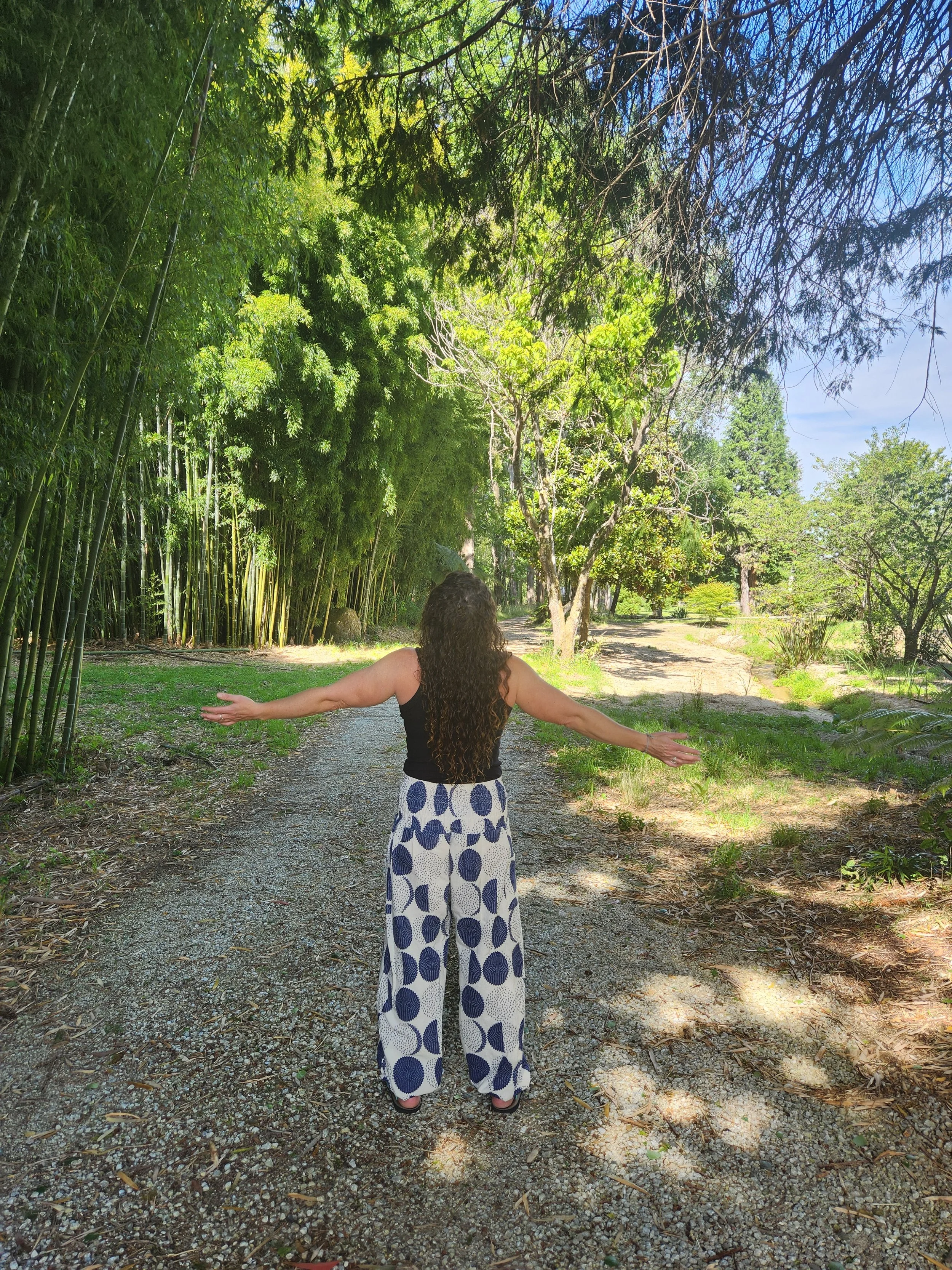 A woman with long curly hair, wearing a black top and wide white pants with blue polka dots, stands on a gravel path with arms outstretched in a lush, green outdoor setting.