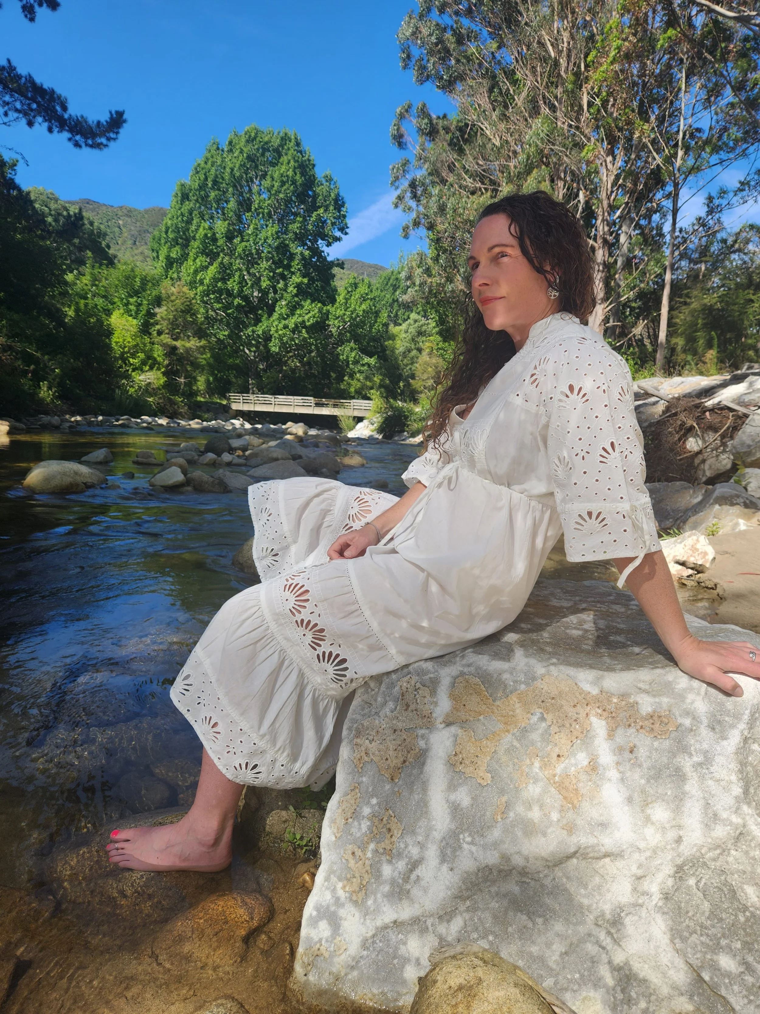 A woman in a white dress with eyelet embroidery sitting barefoot on a large rock by a flowing river, surrounded by lush green trees and a clear blue sky.