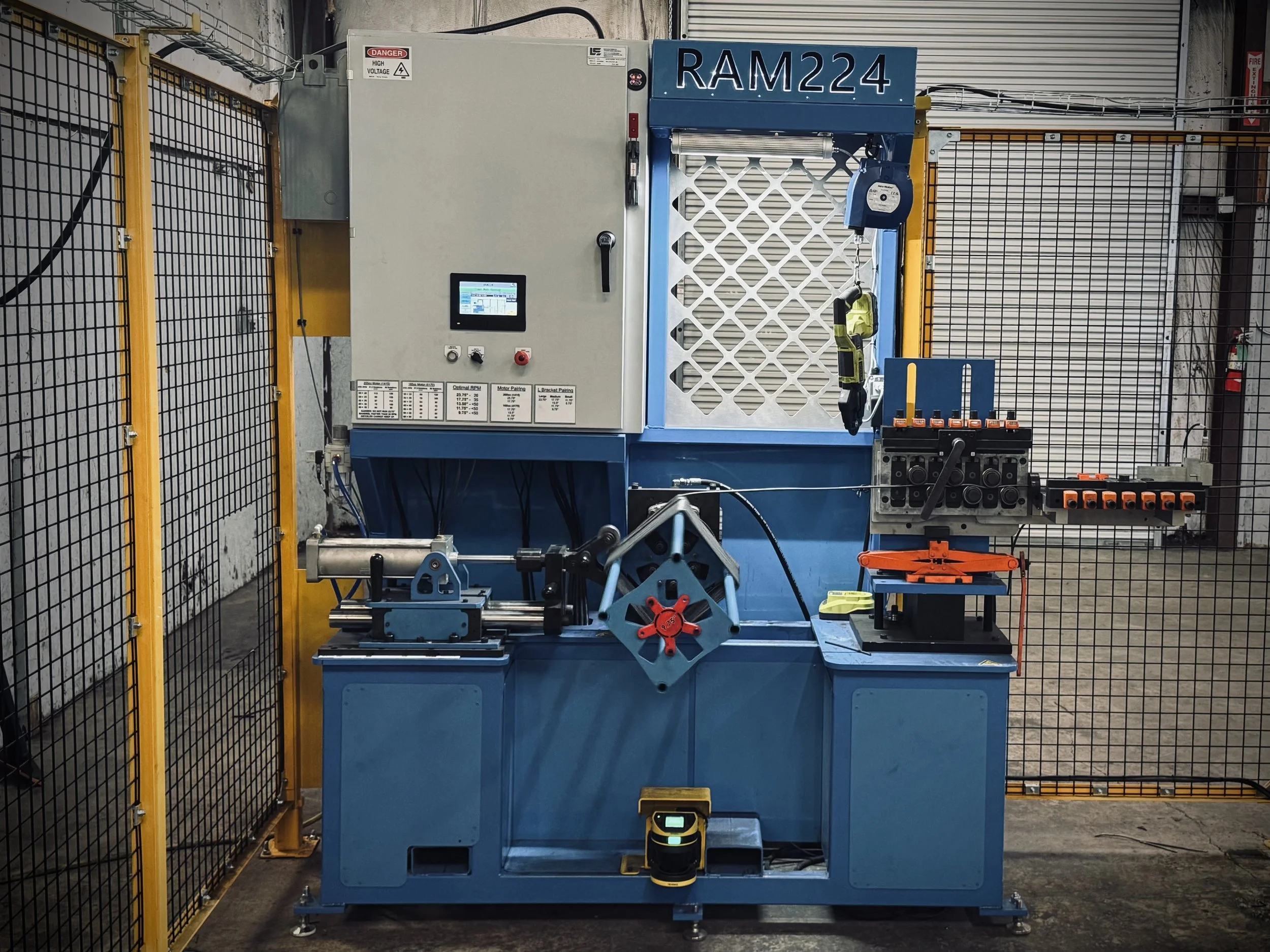 Industrial machine labeled RAM224 with control panel, safety fencing, and tools attached, situated in a warehouse or manufacturing setting.