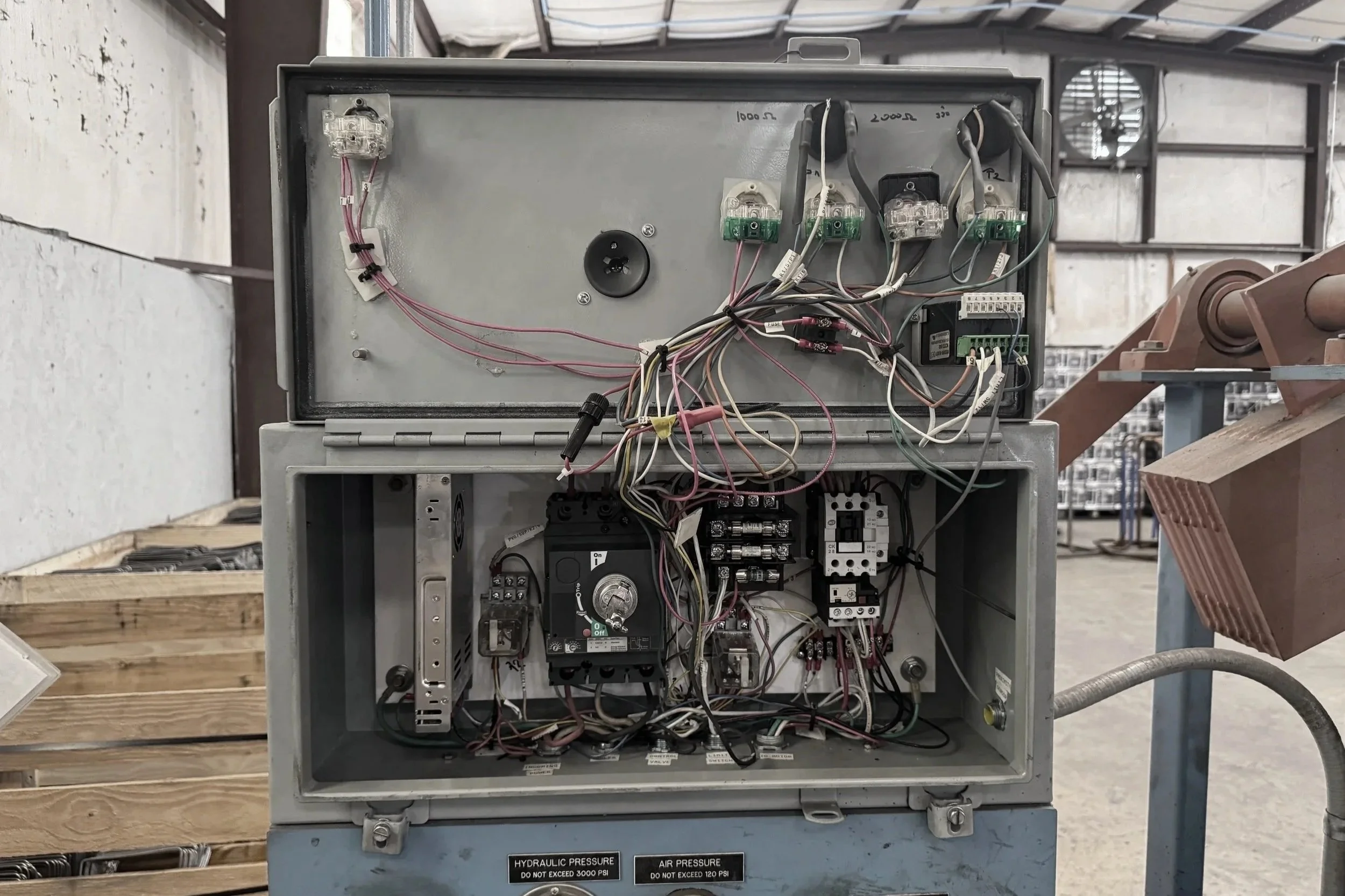 Open electrical control box with wiring and components inside on a workbench in a workshop.