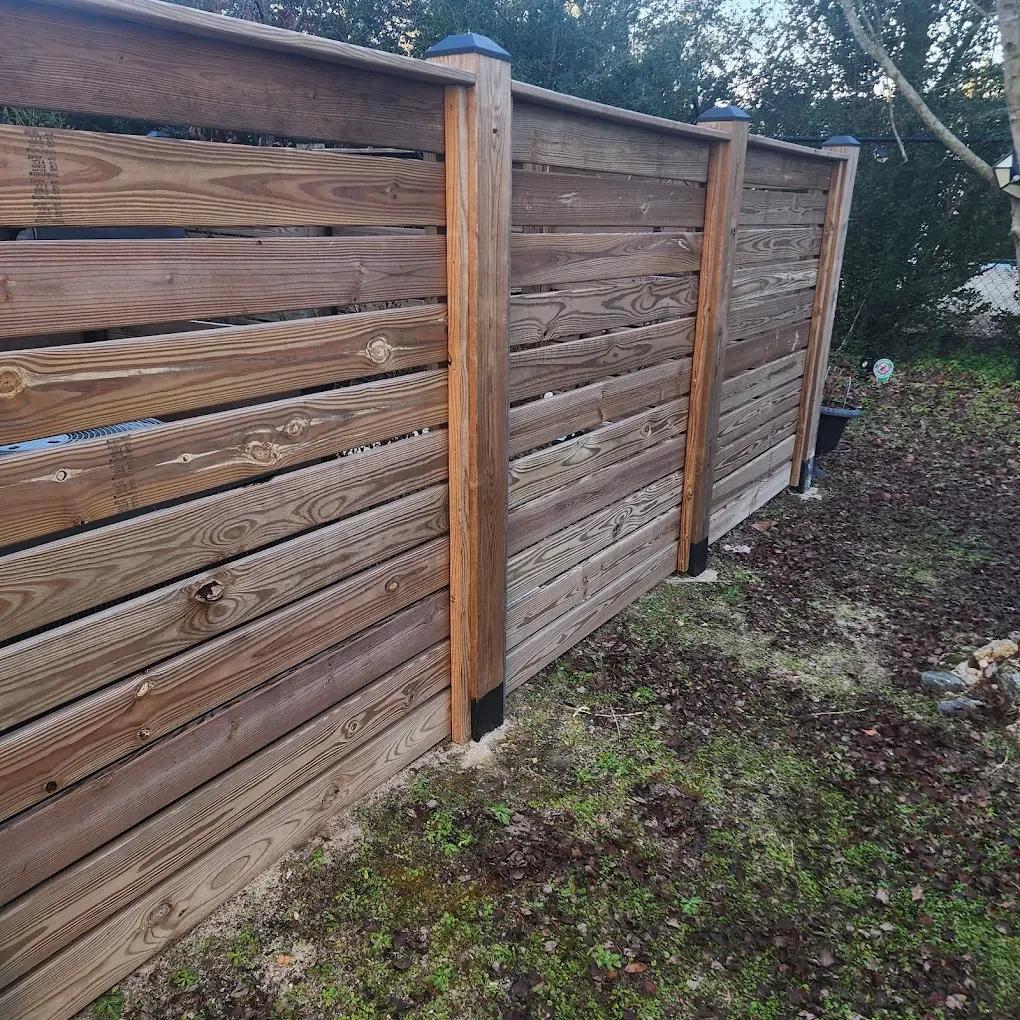 A newly installed wooden privacy fence with horizontal slats and vertical support posts in a yard with dirt and small green plants.