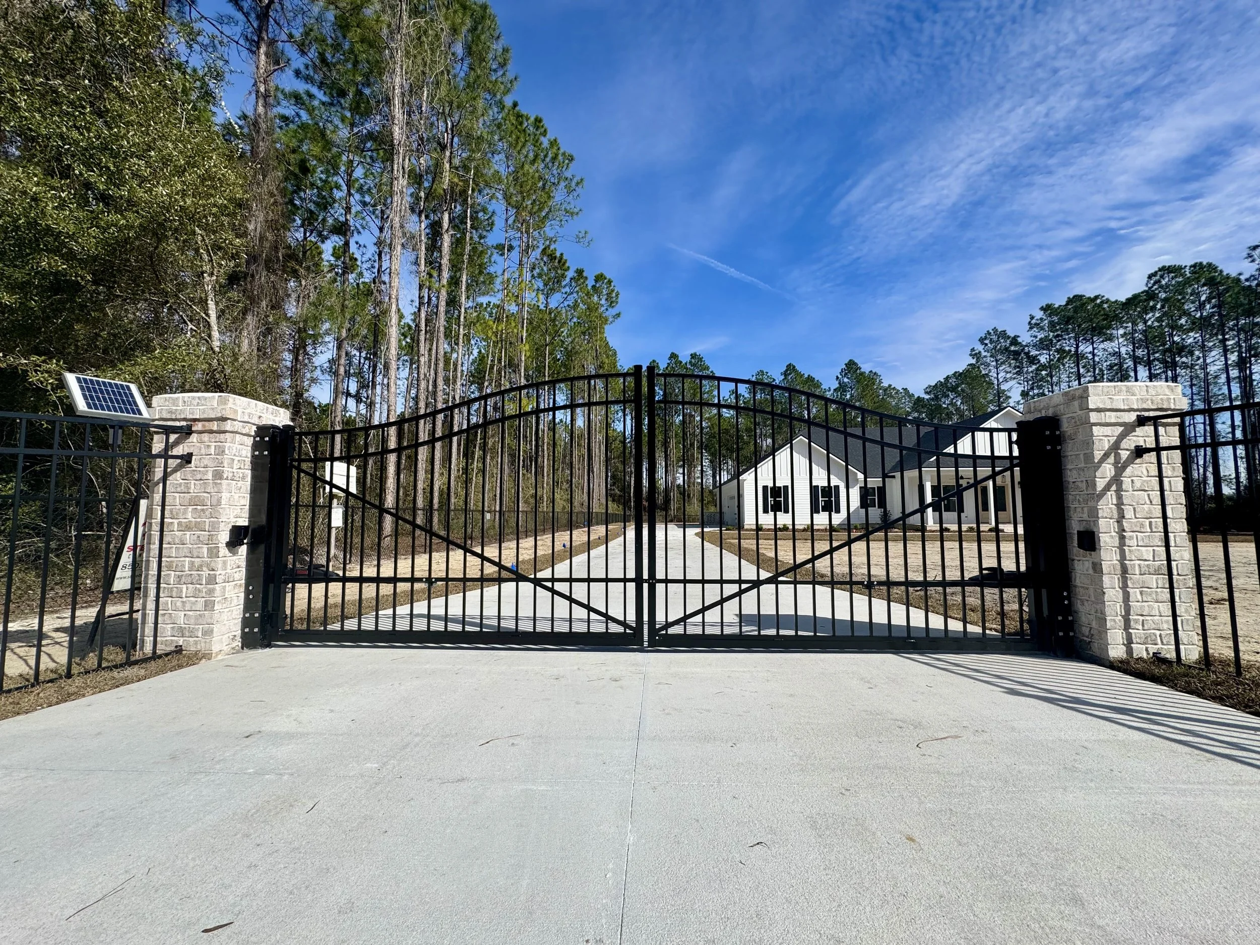 A black metal gate with a curved top and brick pillars at each side. Behind the gate, a white house with black shutters is visible, surrounded by a dirt yard. Tall pine trees are in the background under a blue sky with some clouds.