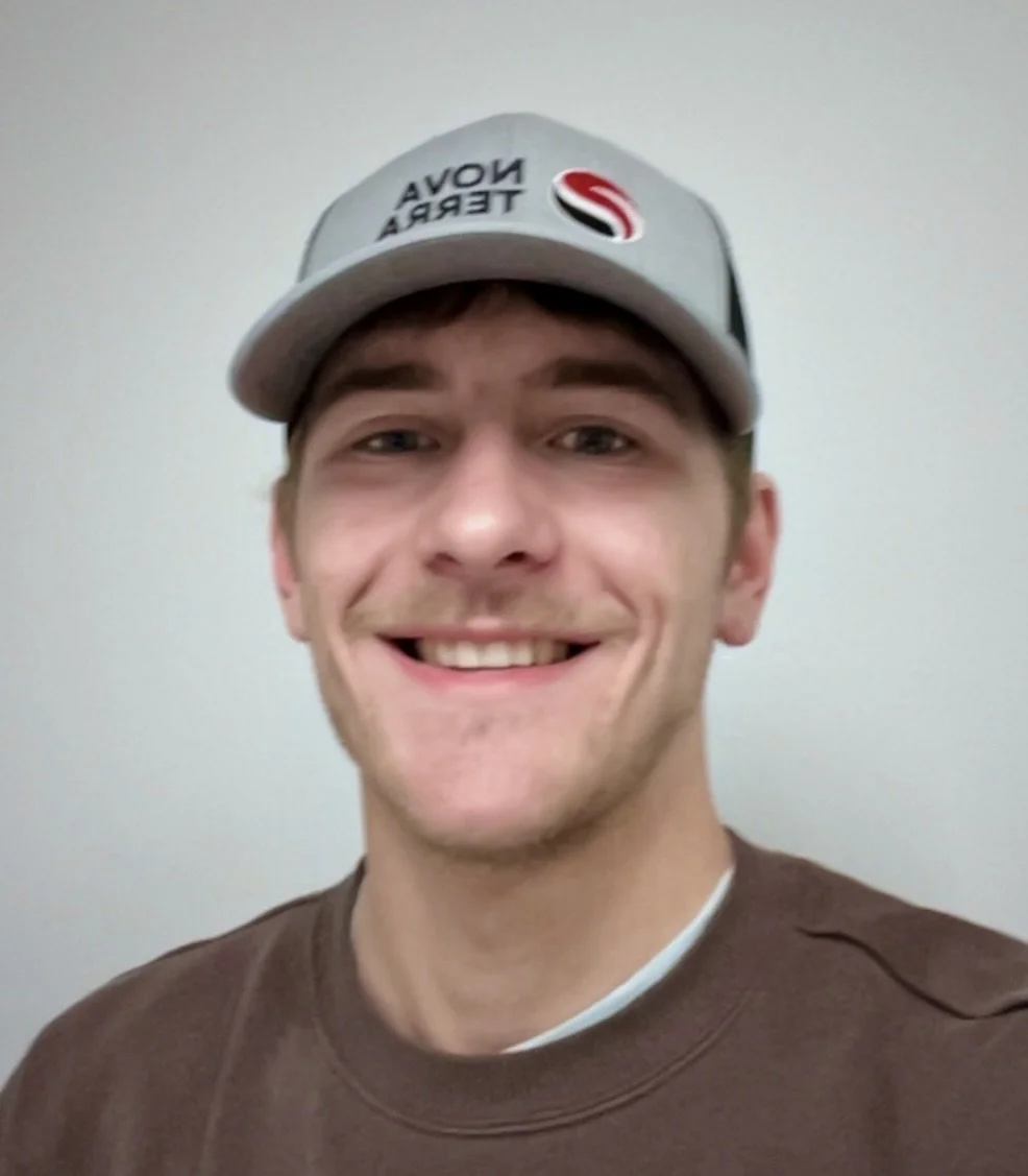 Young man smiling wearing a gray baseball cap with the words 'NOVIA TERA' and a logo, and a brown shirt, standing against a plain light-colored wall.
