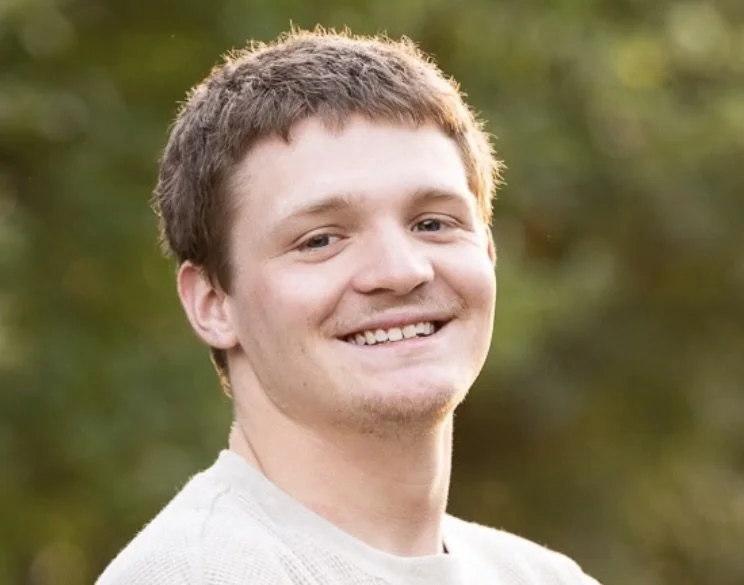 A young man with short brown hair smiling outdoors with a blurred green background.