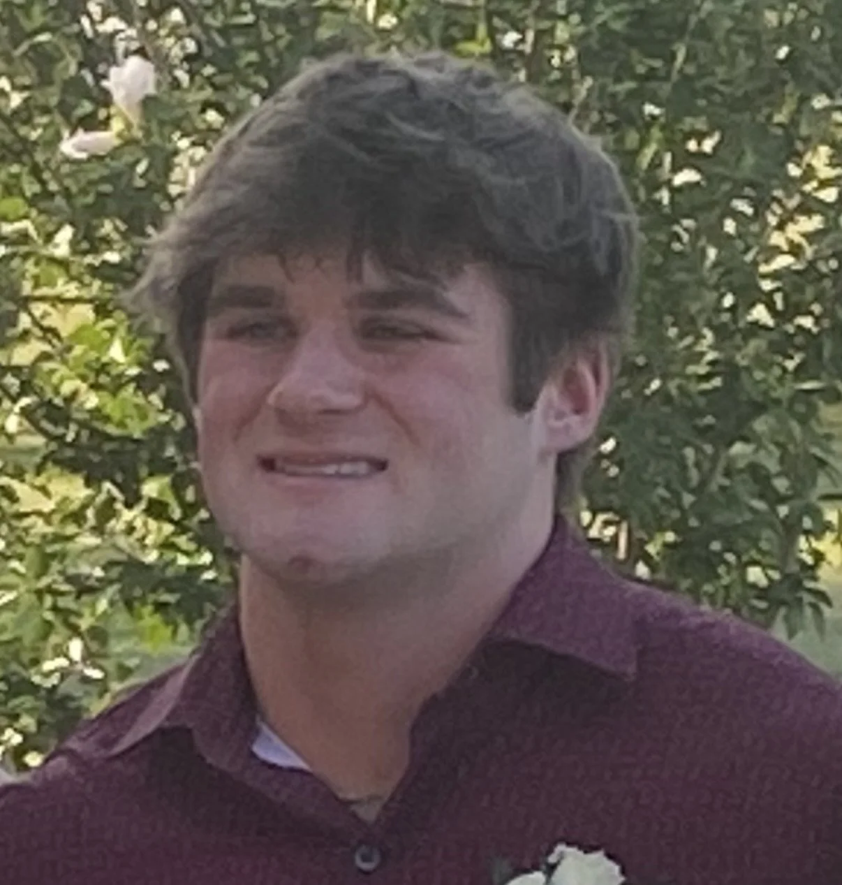 Young man with curly hair smiling outdoors with green leafy background.