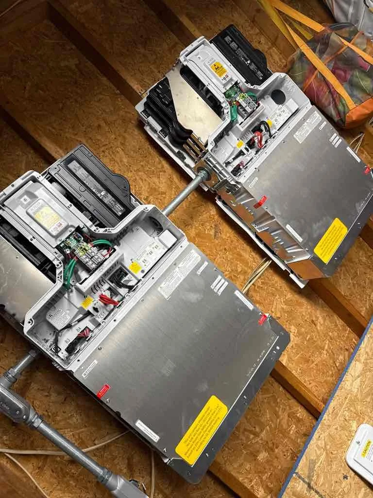 Two large electronic devices with exposed wiring and internal components, connected by metal pipes and set on a wooden surface.