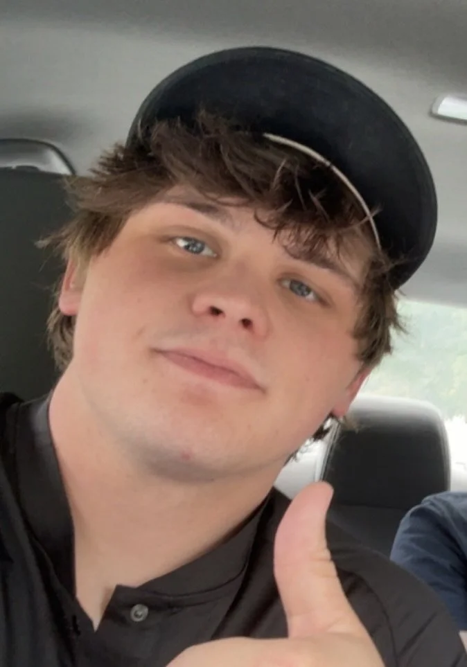 A young man with light skin, brown hair, and blue eyes giving a thumbs up in a vehicle, wearing a black cap and a black shirt.