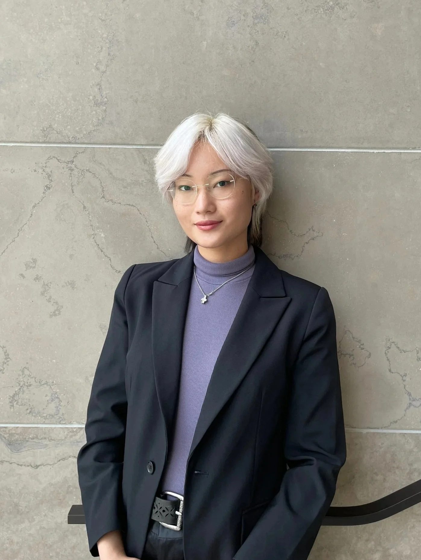 A woman with short, white and purple hair, wearing glasses, a black blazer, and a purple turtleneck, standing against a beige stone wall.
