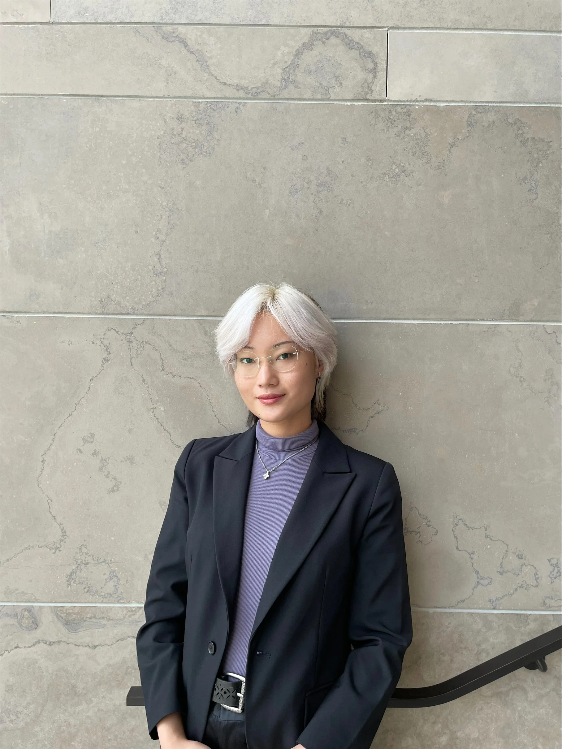 A woman with short, light-colored hair, wearing glasses, a black blazer, and a purple turtleneck, standing against a textured light gray wall.