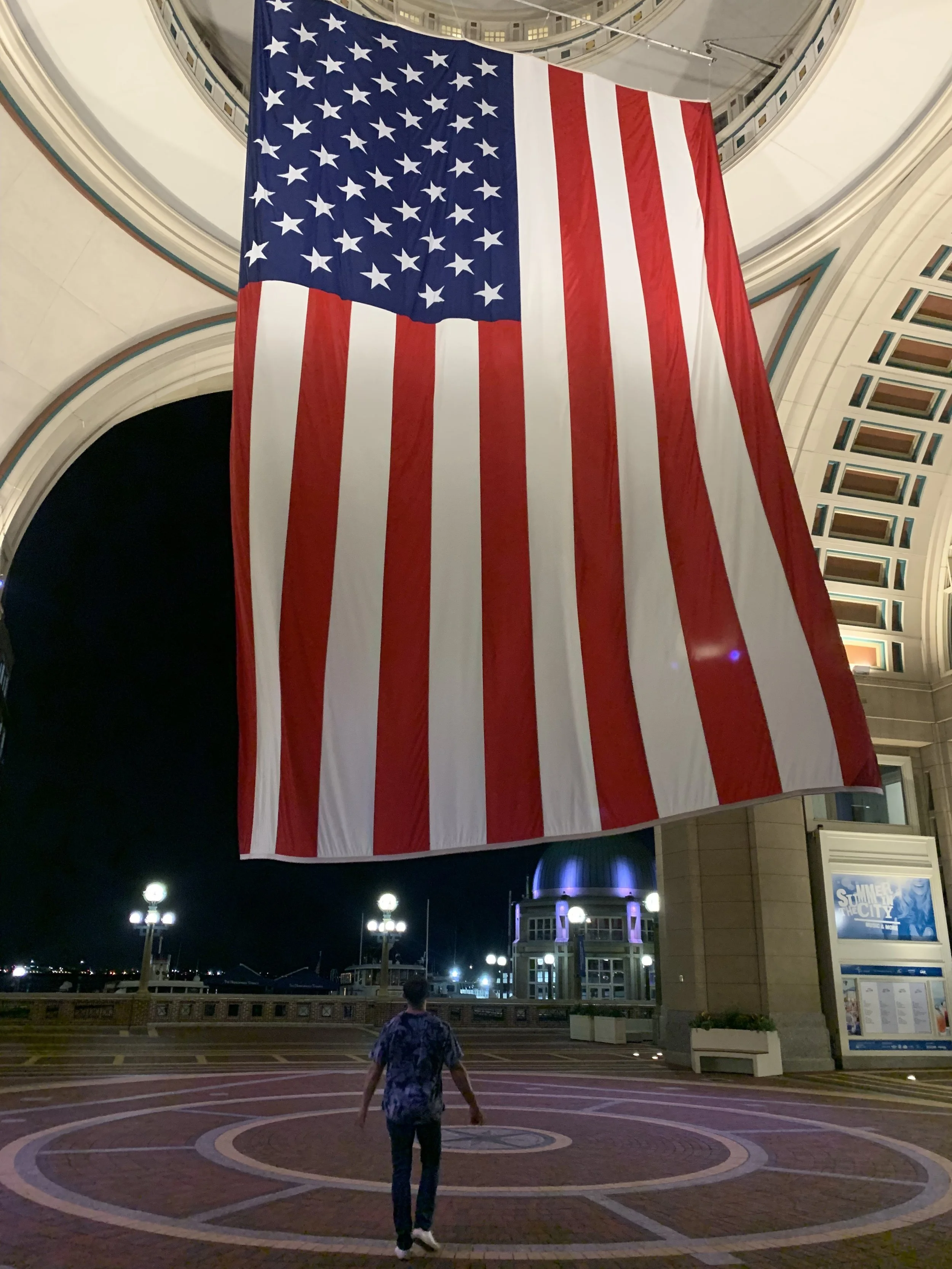 A large American flag hanging indoors over an open space with a person walking underneath at night. The area has decorative lighting and a lit dome structure in the background.