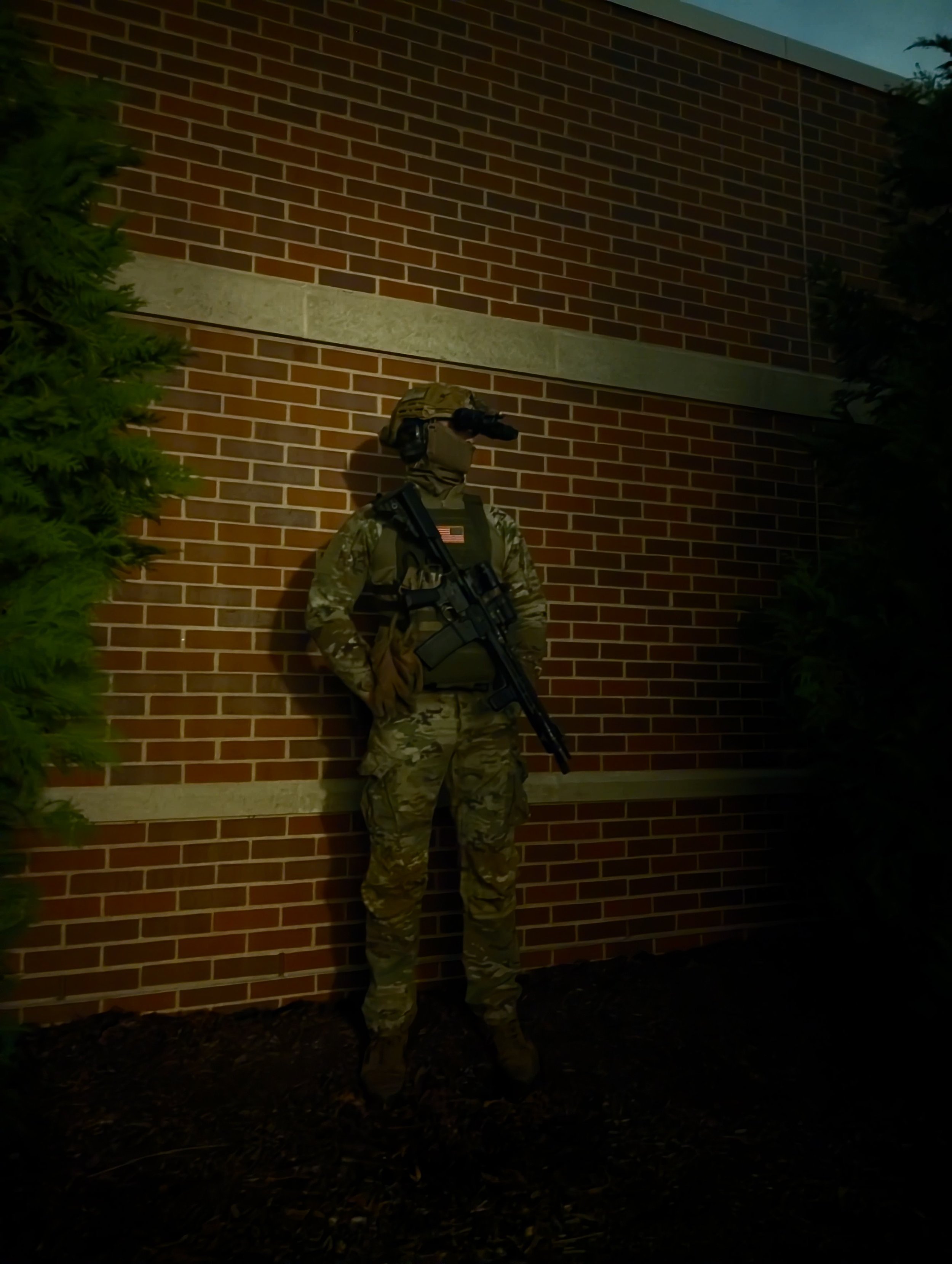 A soldier in camouflage uniform and helmet standing against a brick wall at dusk or dawn, holding a rifle.