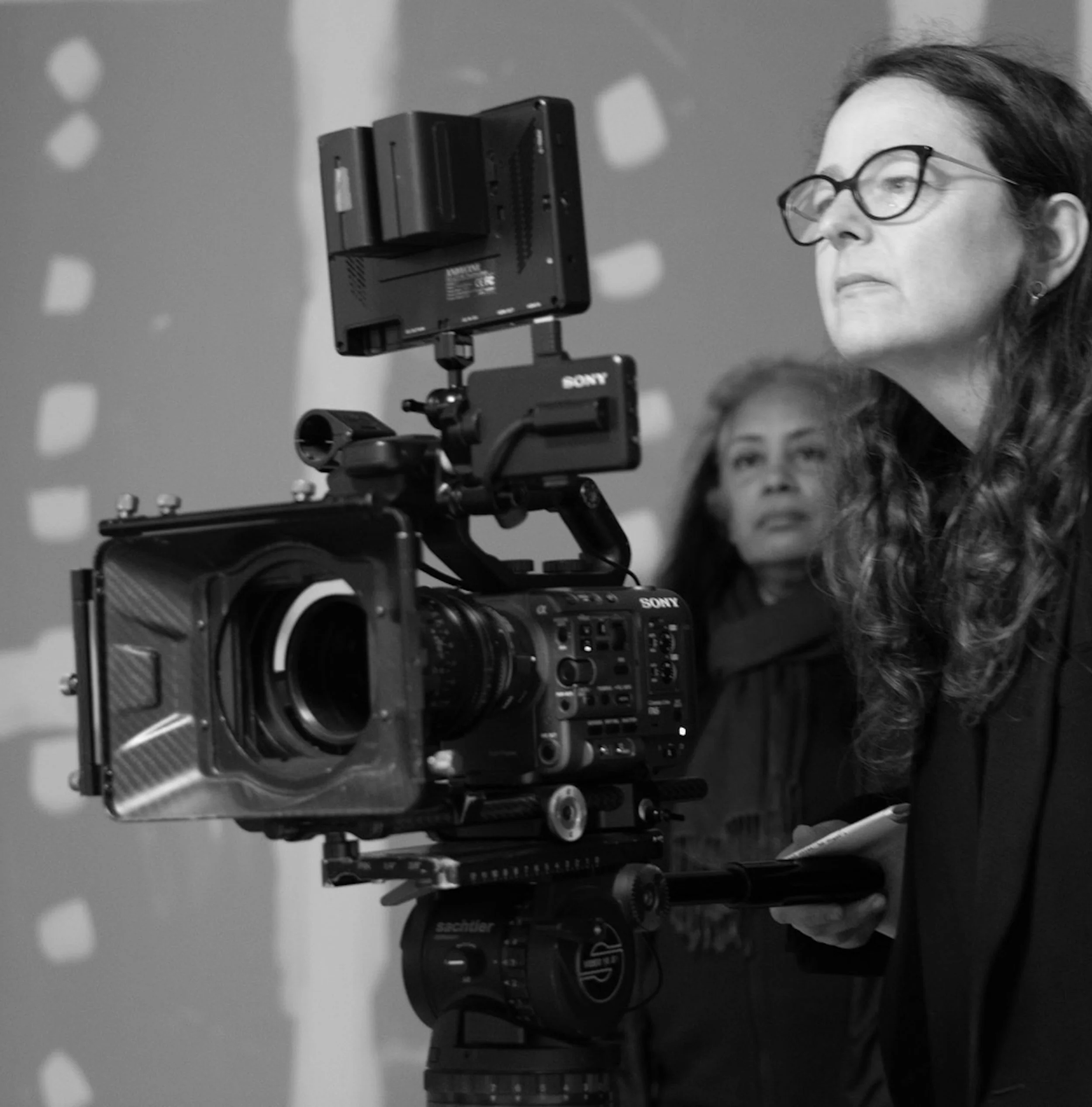 A woman with glasses operating a professional video camera on a tripod, with another woman watching in the background.