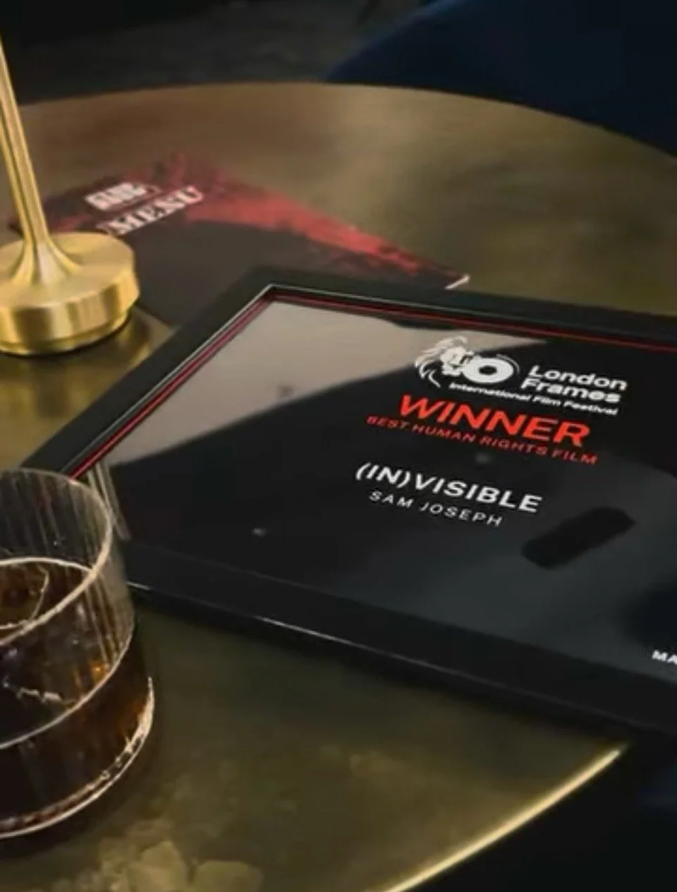 A framed award from the London Frigh M Festival titled 'Winner' for the documentary '(In)Visible' by Sam Joseph, placed on a table next to a drink in a glass and part of a magazine with red accents.