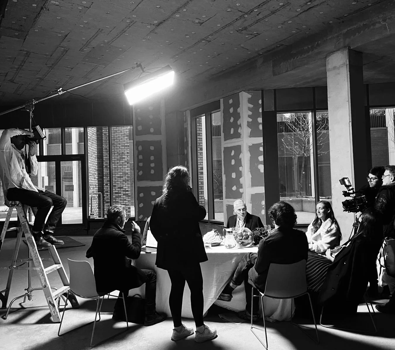 A black and white photo of people in a room with large windows, some seated at a table and others standing or filming, during what appears to be a recording or interview session.