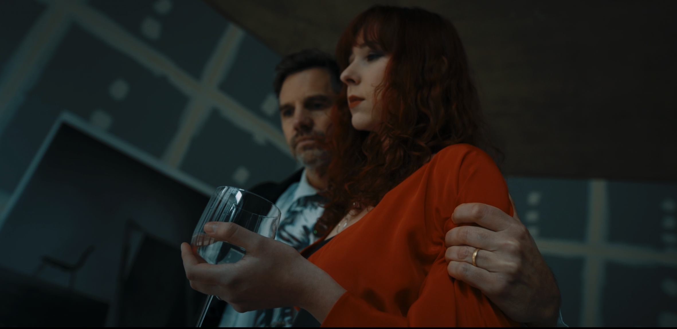 A man and woman in an office setting with a modern interior, the woman in a bright orange dress holding a glass of water while the man, dressed in a shirt and blazer, gently supports her arm.