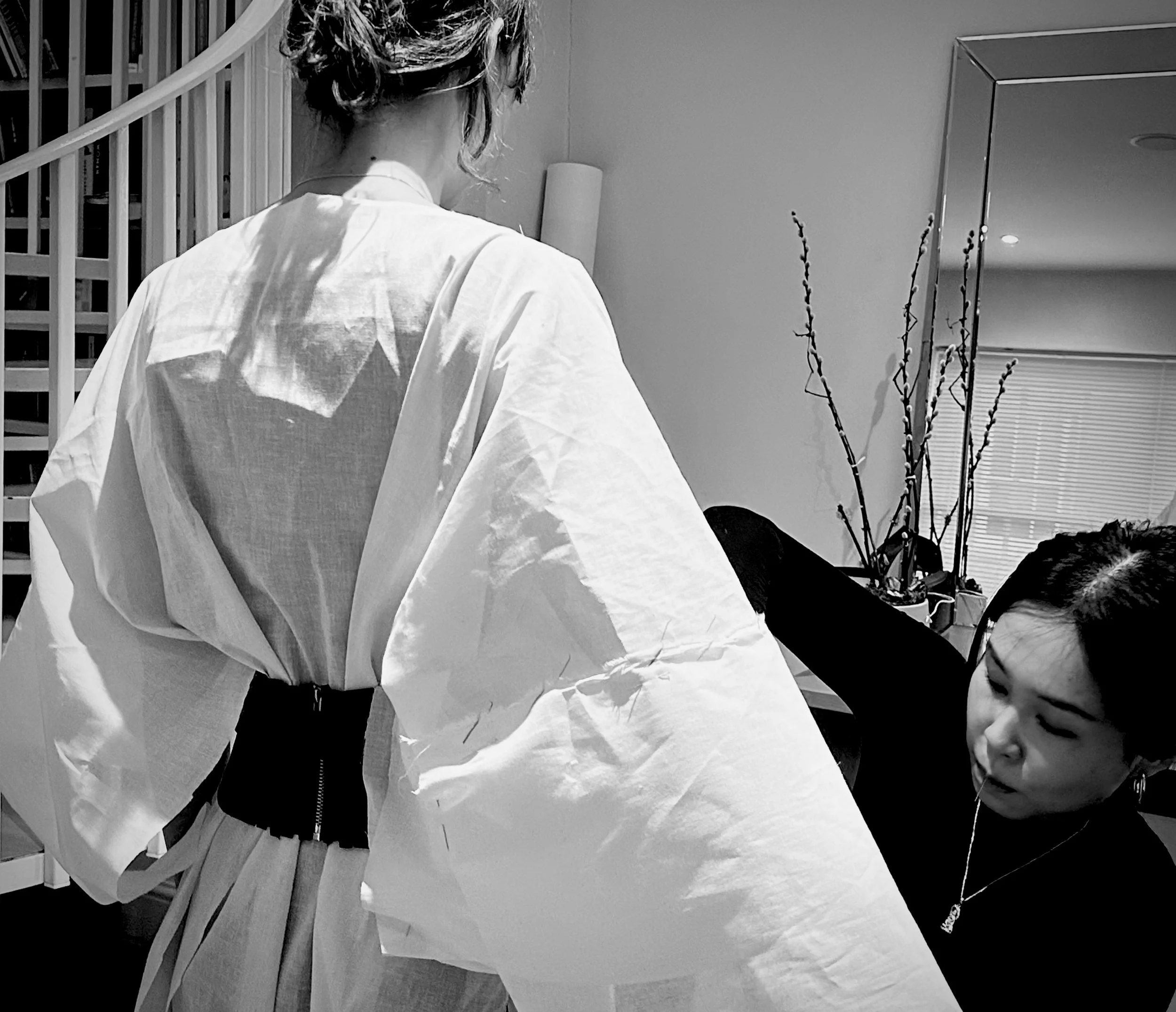 Two women in a room, one standing with her back to the camera wearing a loose white shirt, the other sitting and adjusting her outfit, with a mirror, window blinds, and a plant in the background.