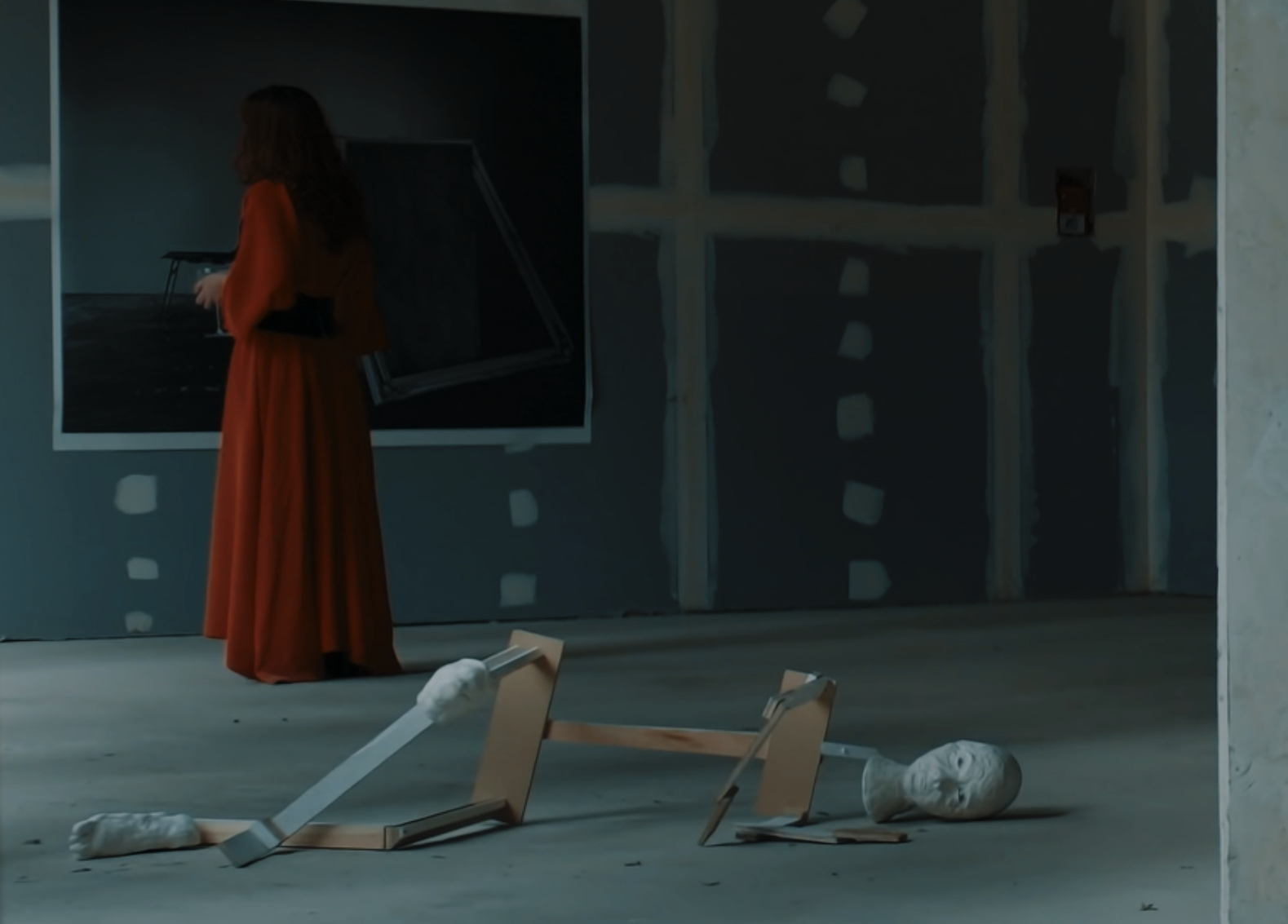 Woman in a red dress standing in an unfinished room, looking at artwork on a wall, with broken wooden frames and a sculpture head on the floor.