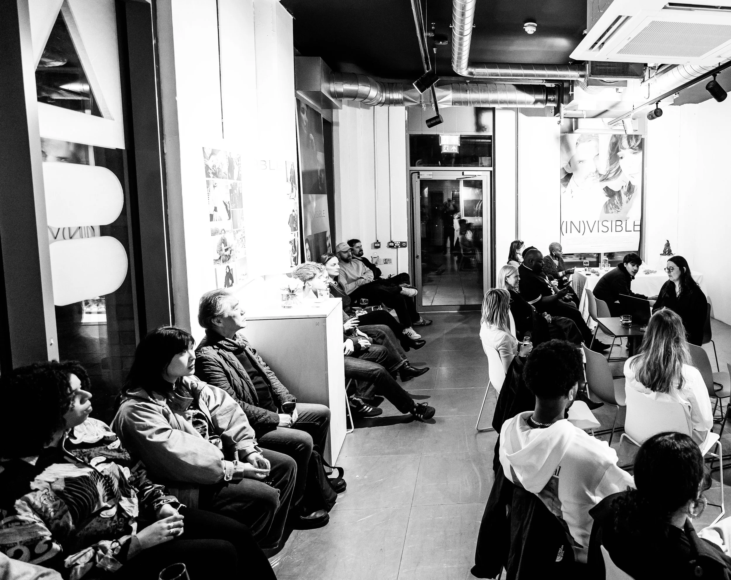 Group of people sitting and listening in a modern indoor space, some with drinks, with a large banner reading 'INVISIBLE' in the background.