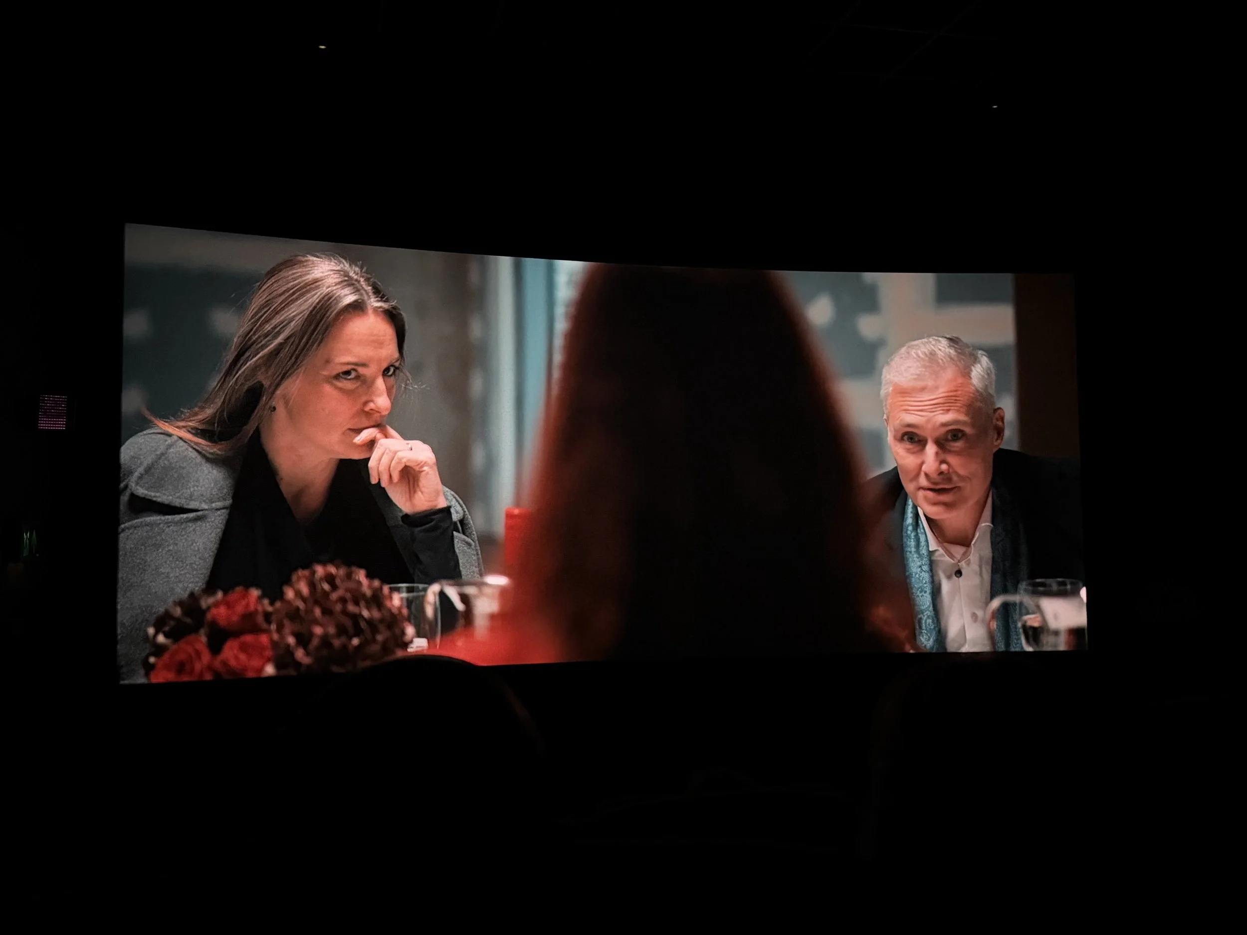 A scene from a movie showing two people at a table, a woman on the left with long brown hair, thinking and resting her chin on her hand, and a man on the right with short gray hair, speaking, with a blurred figure with reddish hair in the foreground.