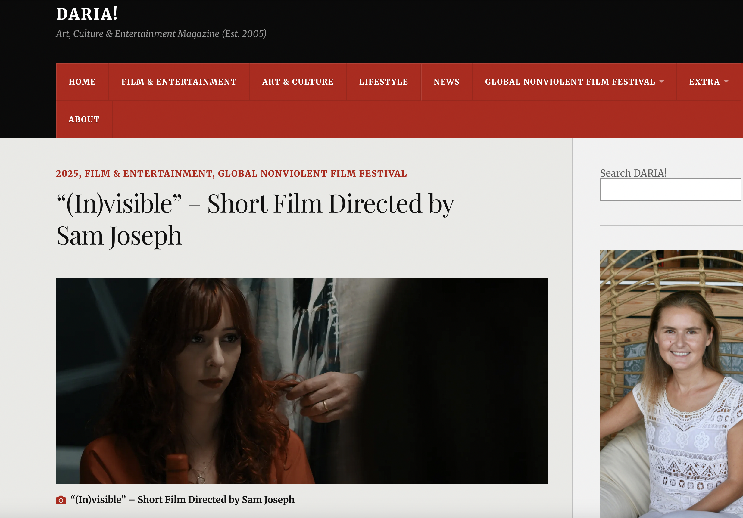 A woman with auburn, curly hair and a serious expression sitting indoors with people around her. The scene appears to be from a film or video set.