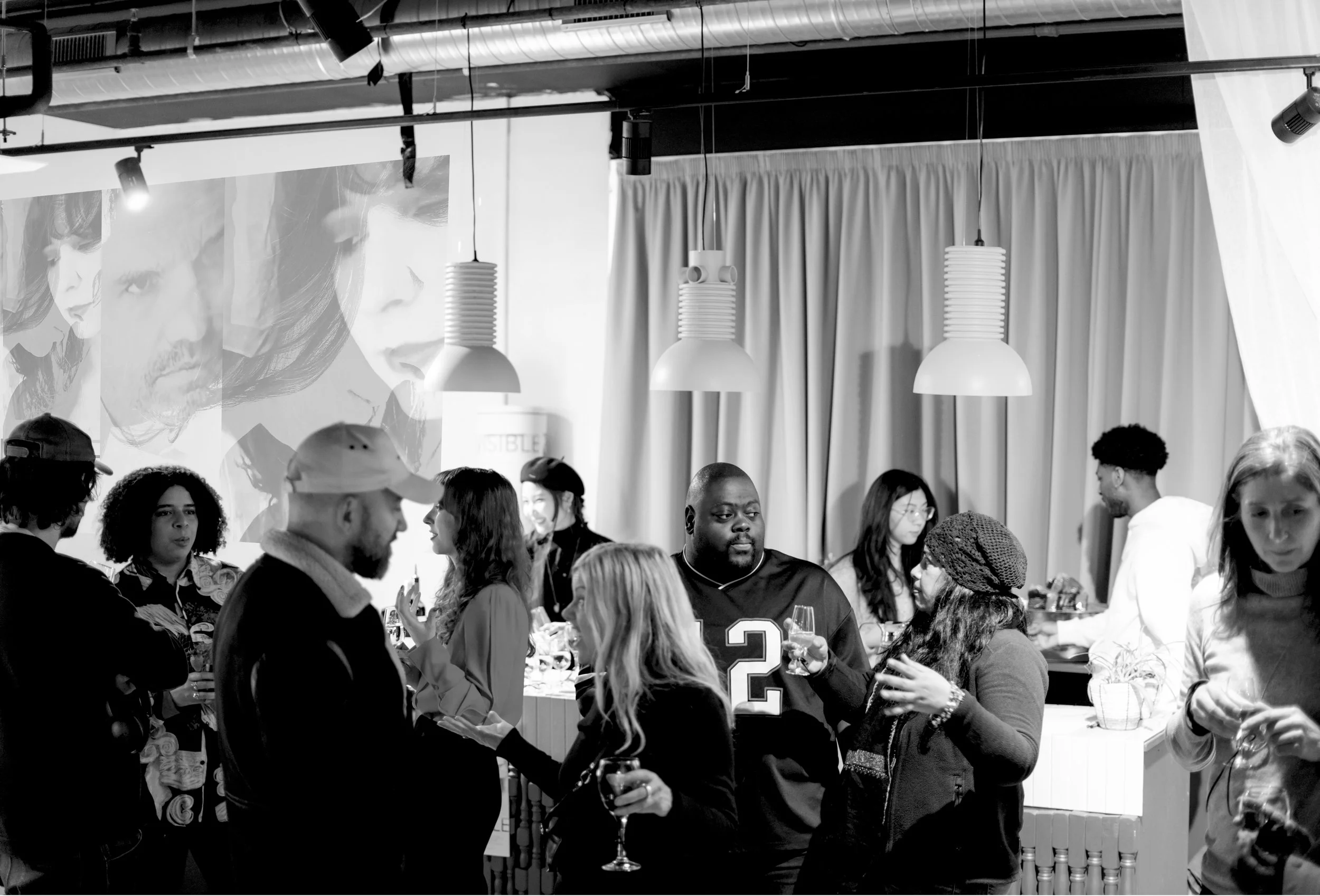 Black and white photo of people socializing at a party indoors, with a large projected image of a woman's face on the wall behind them.
