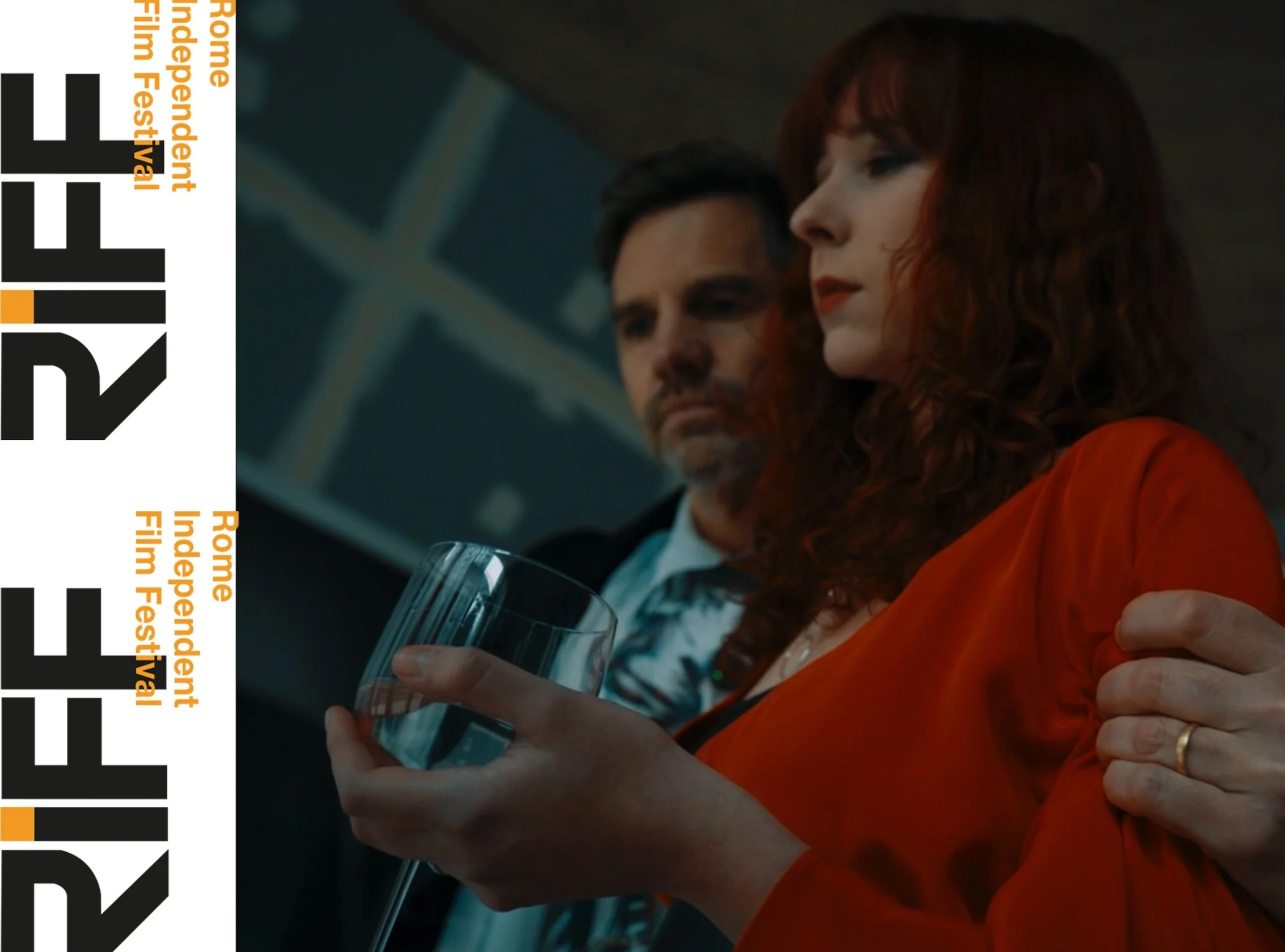 A woman with curly red hair in an orange dress holding a glass of wine, with a man standing behind her, in a dimly lit indoor setting.