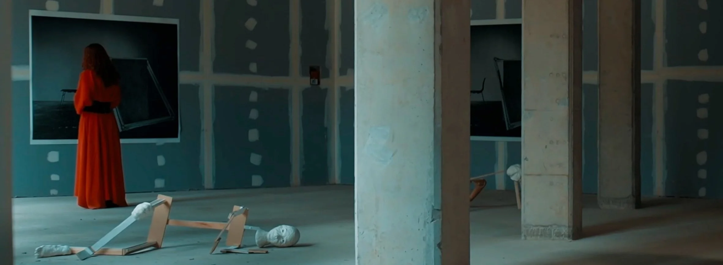 A woman in an orange dress stands in an unfinished room with construction debris, including a broken picture frame and wrapped items, on the floor.