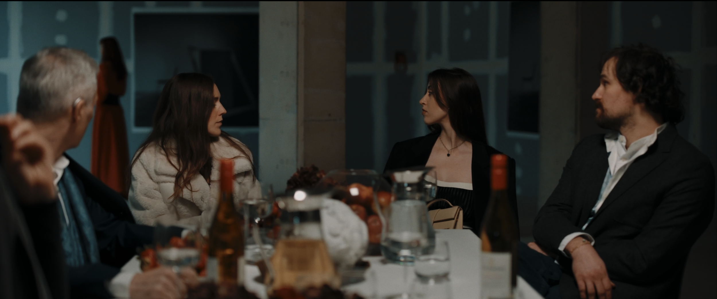 People sitting at a dinner table in a formal setting with food, drinks, and bottles, engaging in conversation.