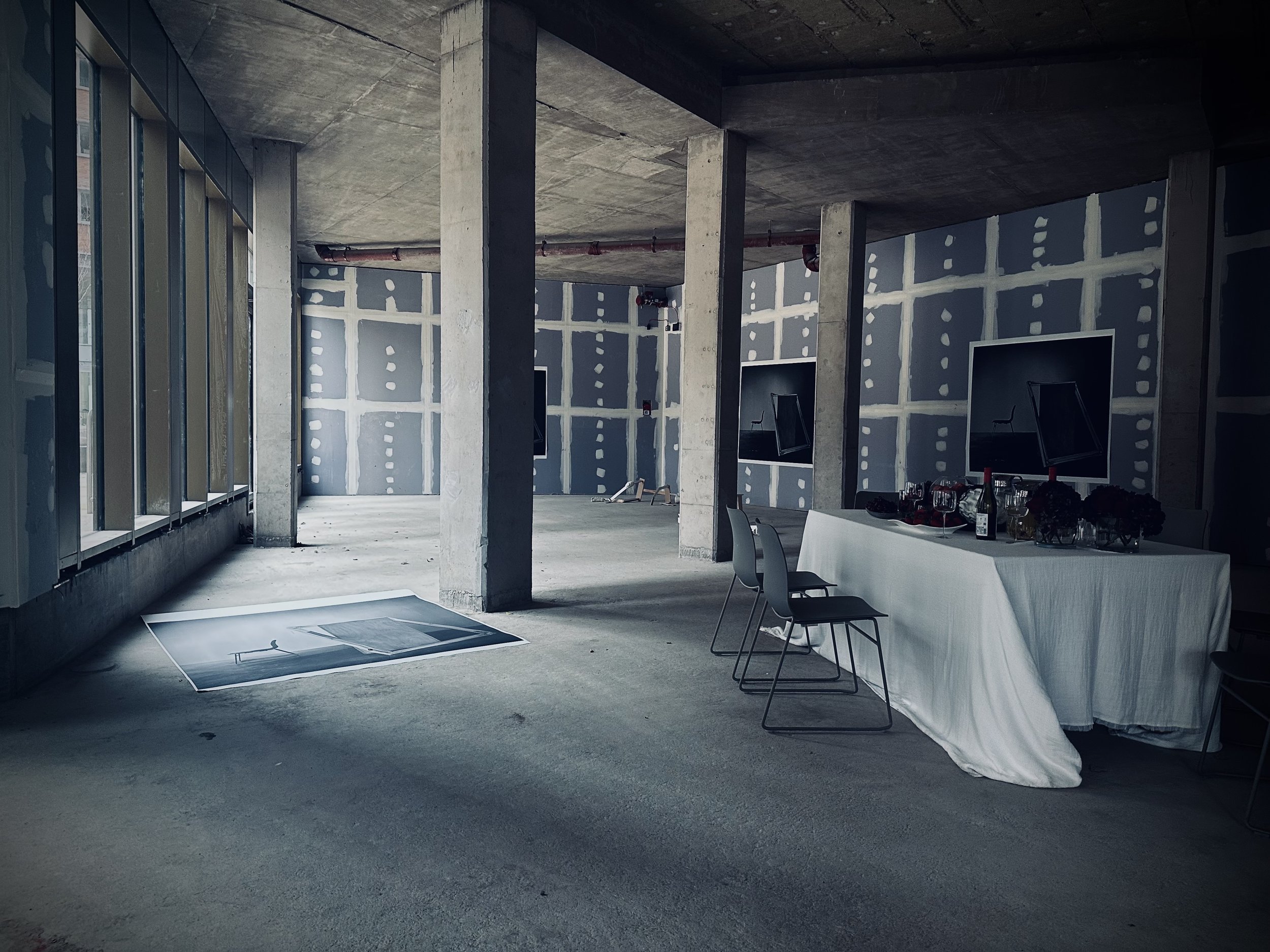 An unfinished interior space with concrete walls and columns, a table with a white tablecloth, chairs, and several monitors on the wall