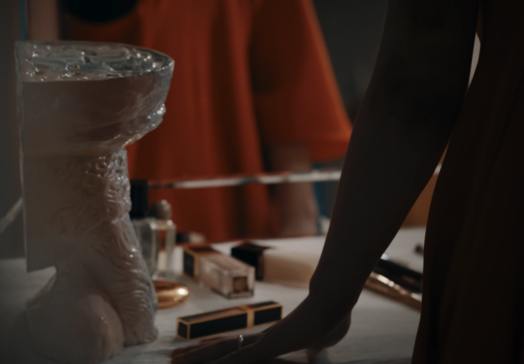 A person in an orange shirt sitting at a table with various objects, including a glass of water, candles, and small rectangular boxes.