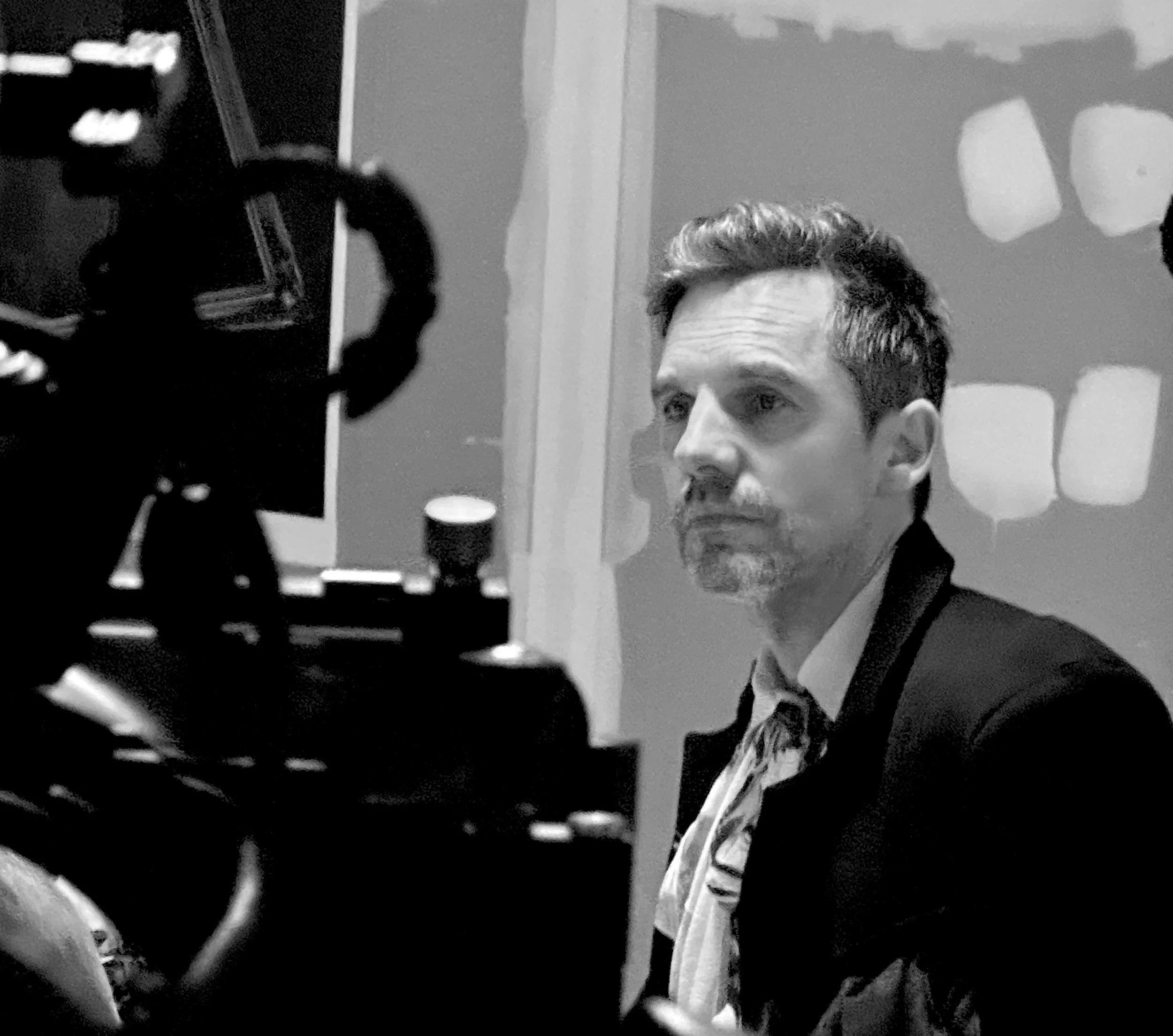 Black and white photo of a man with short, wavy hair and a beard, sitting for a video interview or recording, with professional camera equipment visible in the foreground.