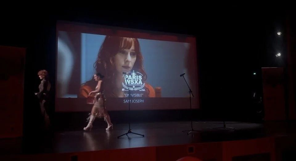 A theater stage with a large screen displaying a woman's face and text about Paris VS SX. Two women are walking across the stage in front of the screen, one standing and the other moving.