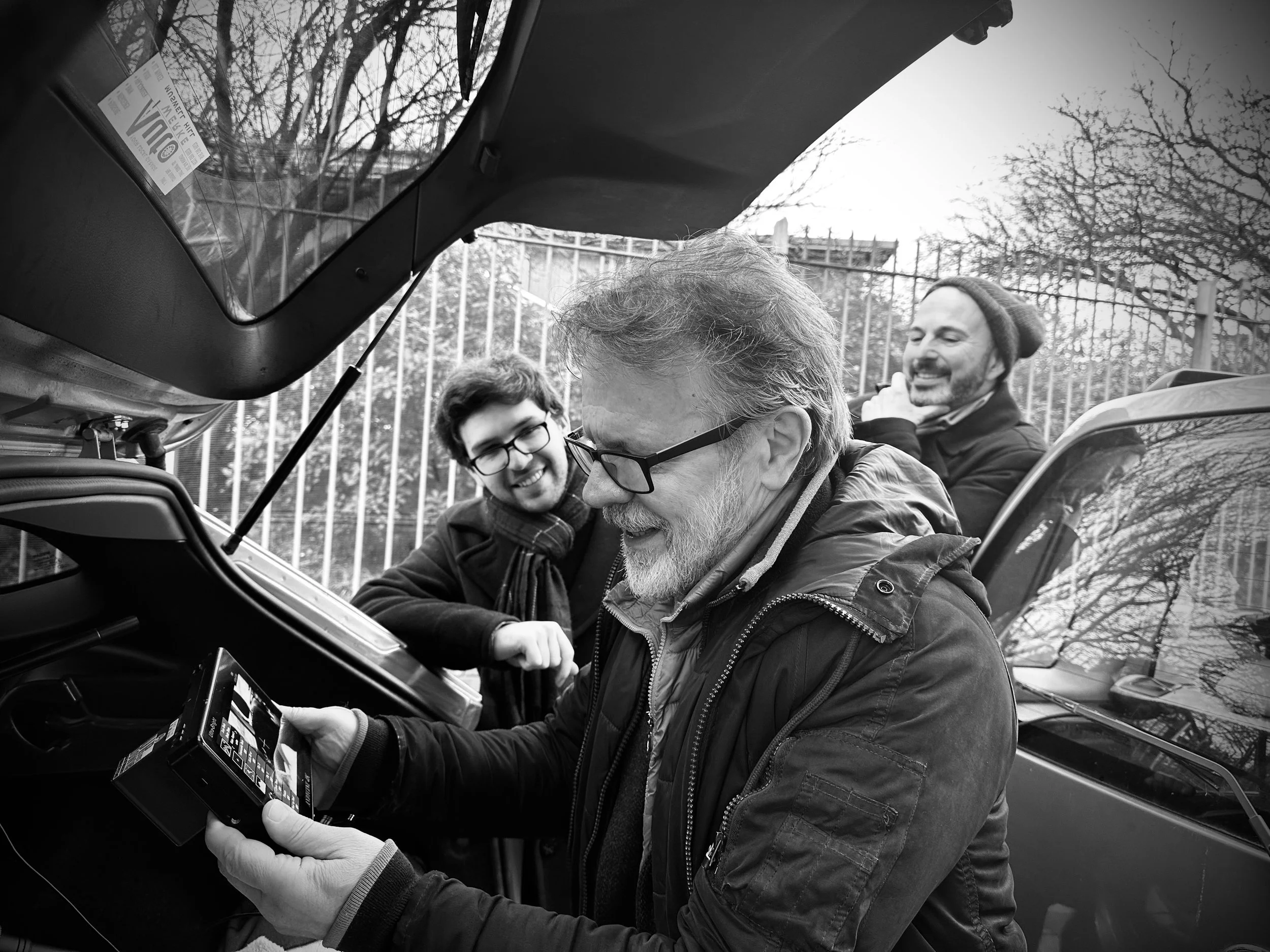 Three men looking at a camera, one man is using it while the other two smile and look on outdoors near parked cars and a fence