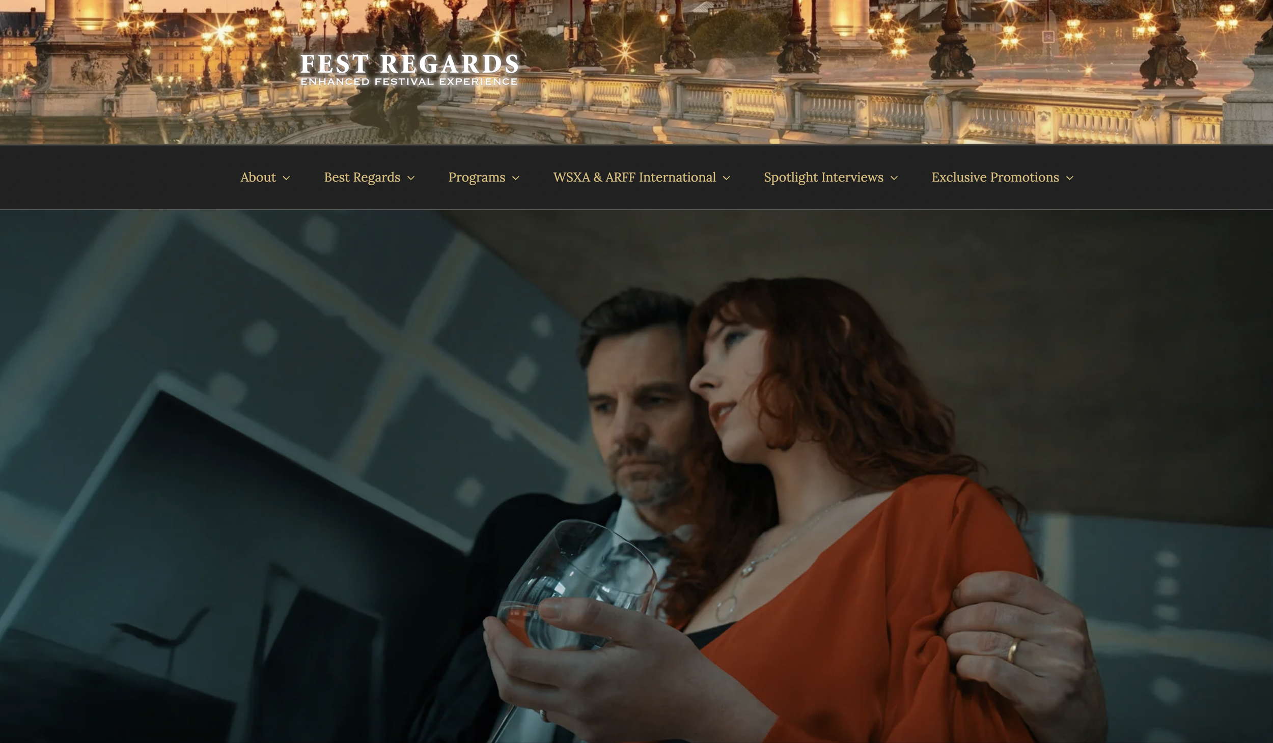 A man and woman dressed formally, with the man gently holding the woman's shoulder, and the woman holding a glass of wine, in an indoor setting with a modern, geometric background.