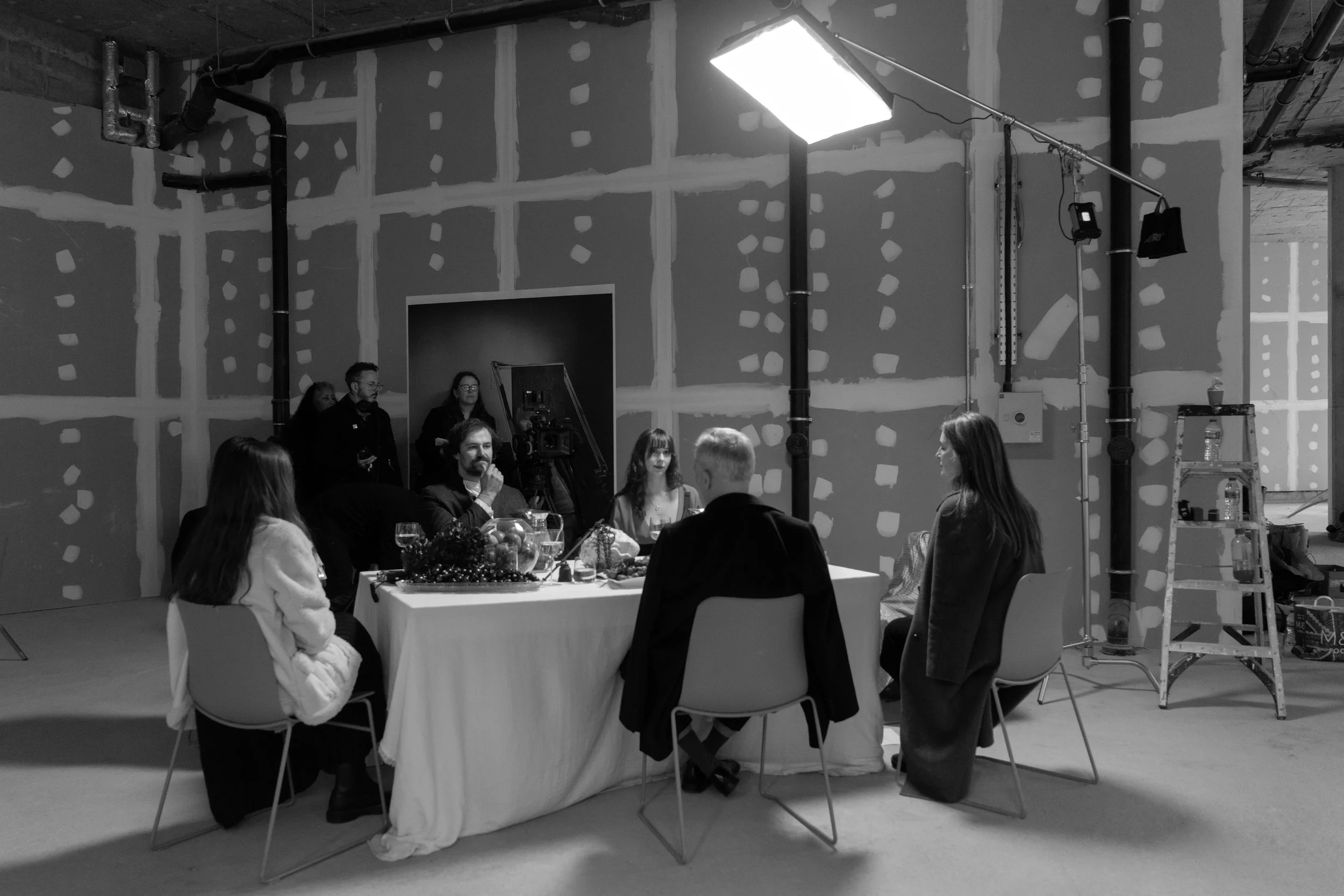 Black and white photo of a group of people sitting around a table, likely filming a discussion or interview, in a studio with unfinished walls and lighting equipment.