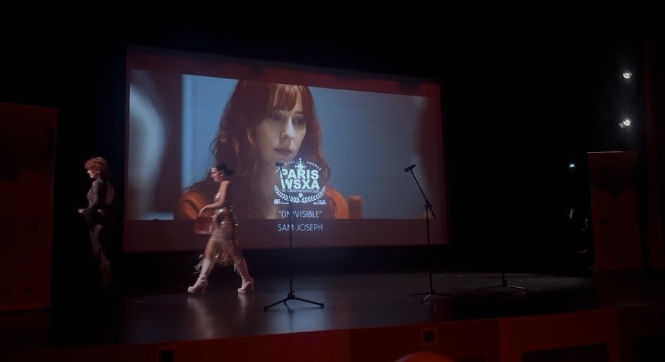 An empty theater stage with a large screen showing a movie scene of a woman with red hair, with the text "PARIS VSXA INVISIBLE" and "SAM JOSEPH".