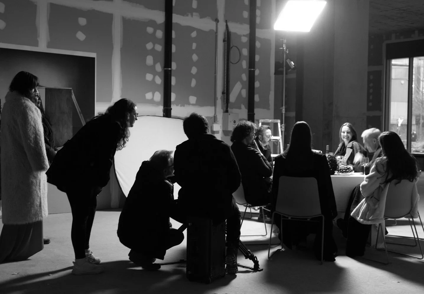 Black and white photo of people gathered around a table in a room with unfinished walls, natural light coming from a window, and studio lighting equipment visible.