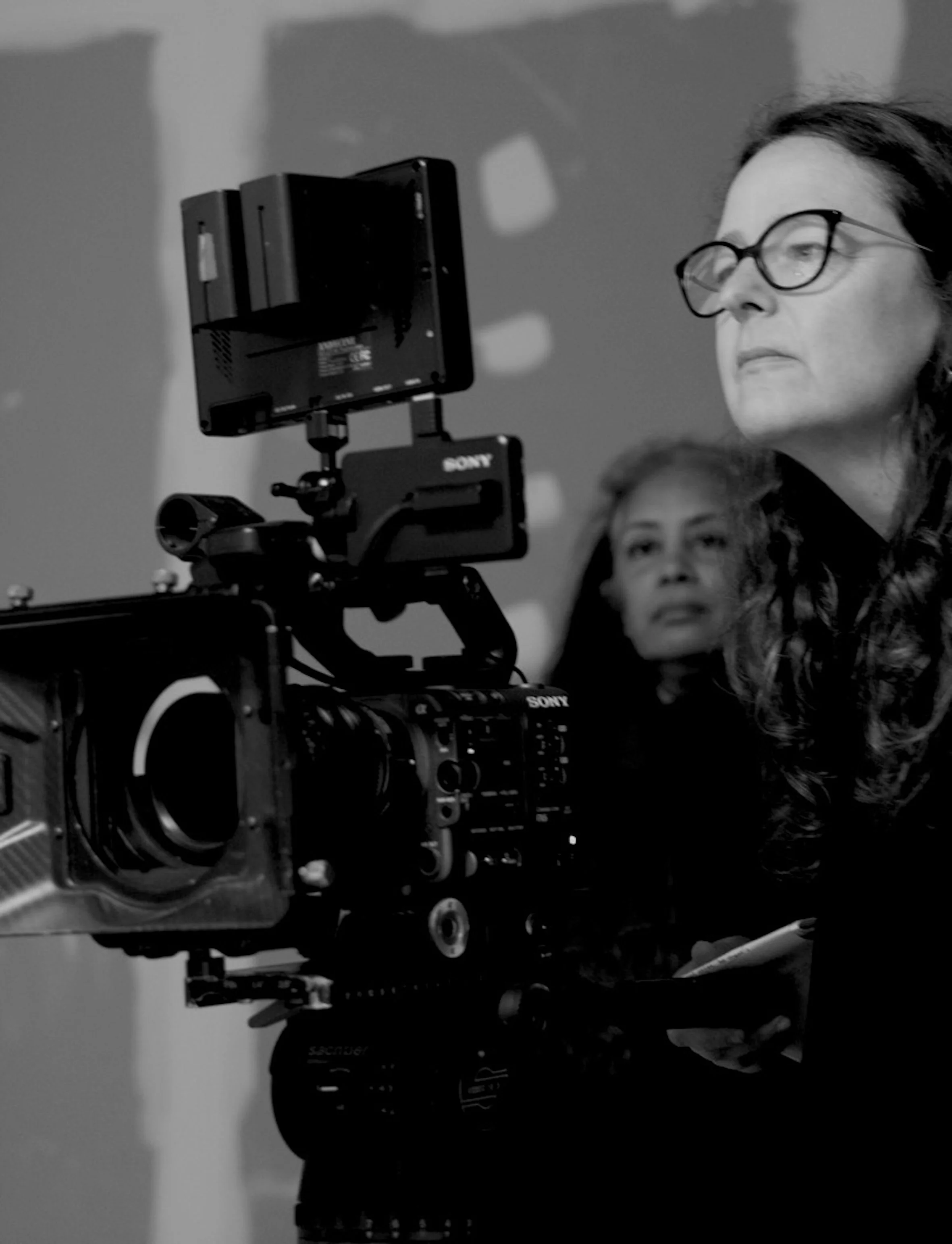 Black and white photo of a woman with glasses operating a professional video camera, with another woman in the background.