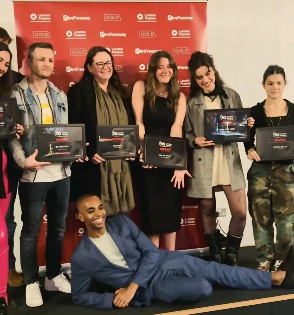 A group of seven young people and one man posing with certificates at an awards event. They are standing in front of a red backdrop with event and sponsor logos, and one man is lying on the floor in front of the group, smiling.