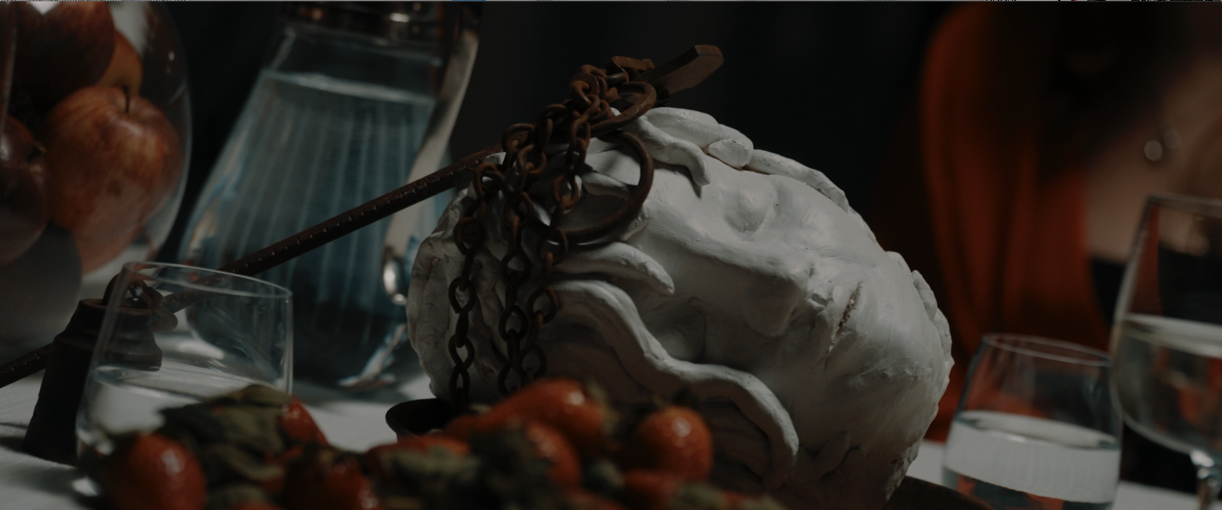 A white sculpture of a human head with a chain and a rusty metal rod draped over it, surrounded by glasses of water, strawberries, and a bowl of apples on a table.