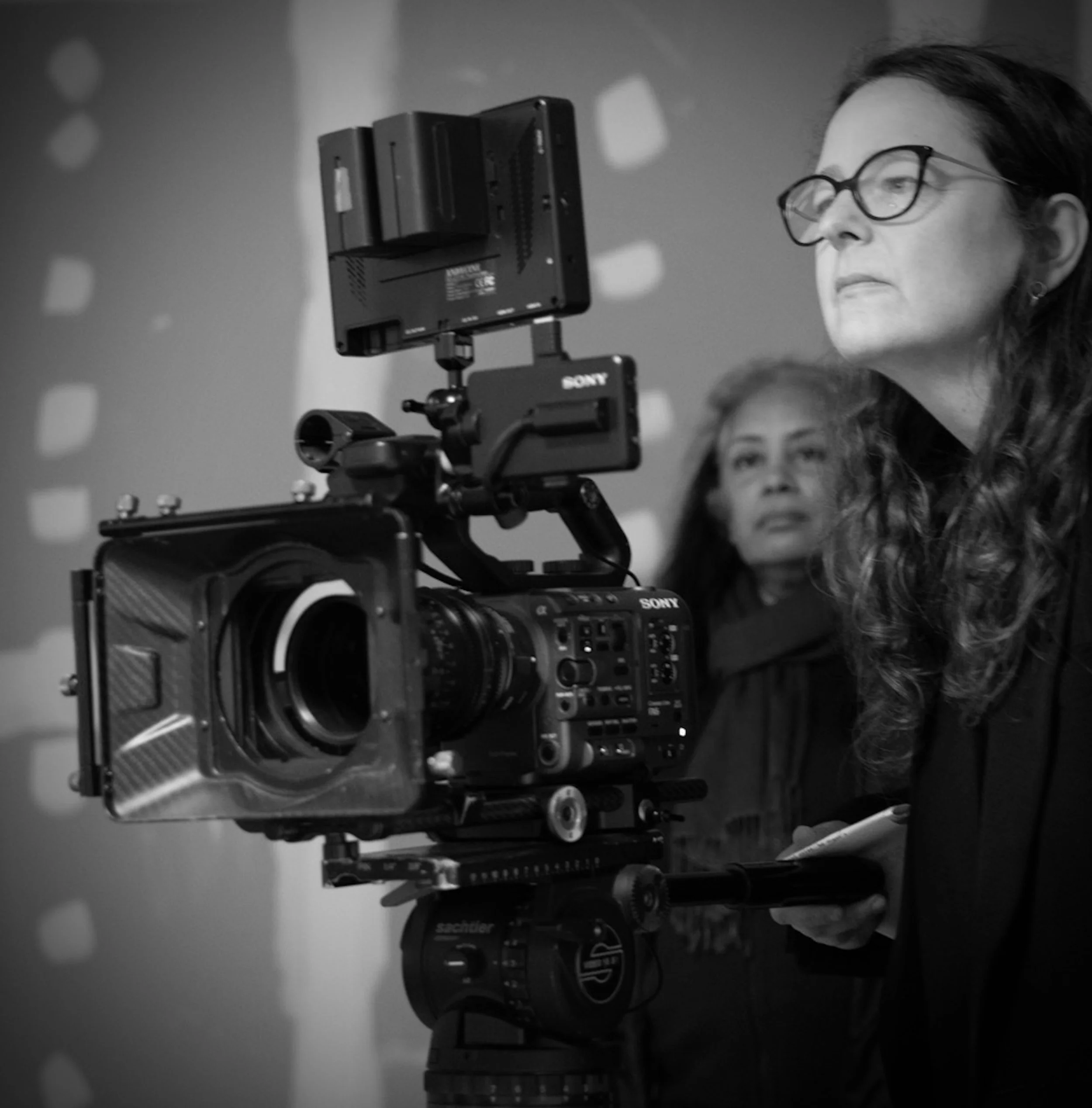 Black and white photo of a woman with glasses operating a professional camera on a tripod, with another woman watching in the background.