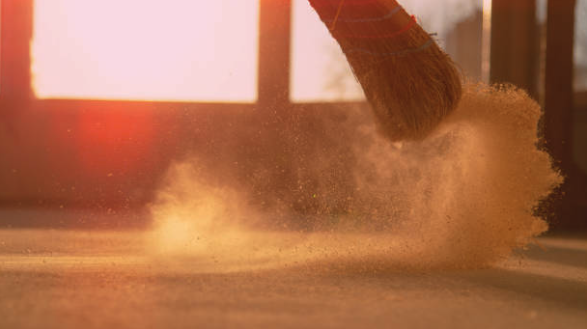 Close-up of a broom sweeping dust on the floor with sunlight in the background.