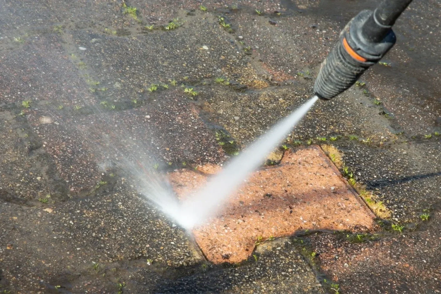 A close-up of a pressure washer spraying water on a brick walkway with small green weeds growing between the bricks.