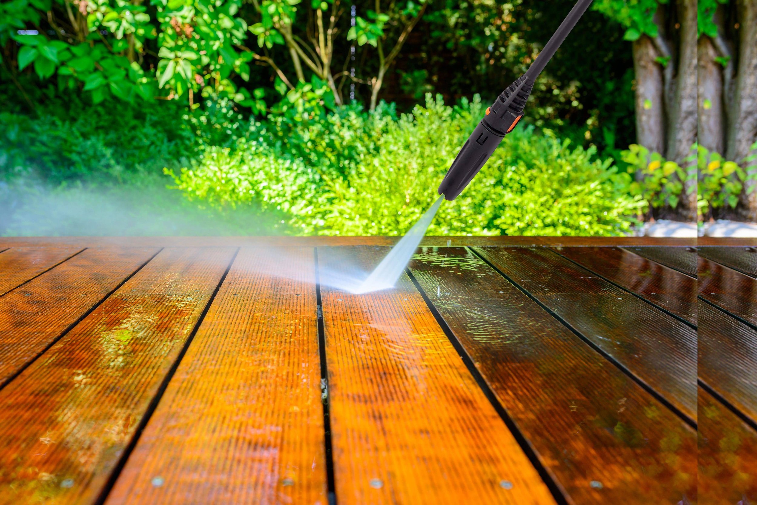 A pressure washer is cleaning a wooden deck outdoors, with green bushes and trees in the background.