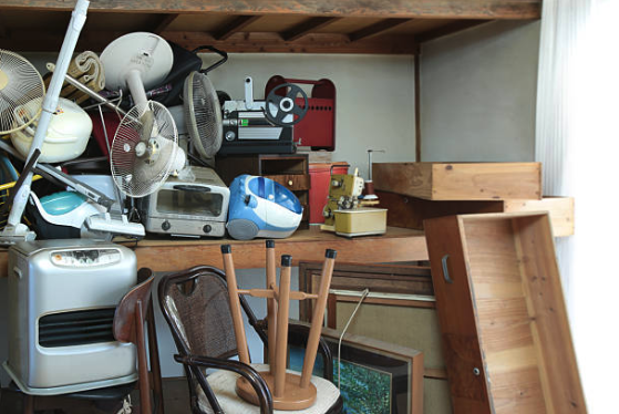 Cluttered room with various electrical appliances, fans, radios, and other objects stacked on shelves and furniture, next to a wooden table and chairs.