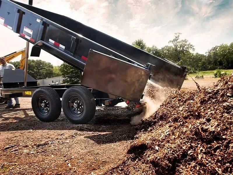 Dump trailer tipping soil or mulch on a dirt surface outdoors under a partly cloudy sky, with trees in the background.