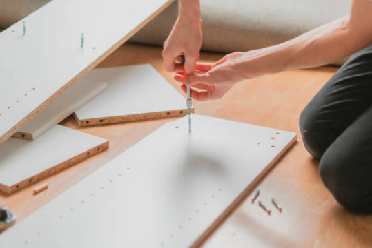 A person assembling furniture by screwing in a bolt into a white board on a wooden floor.