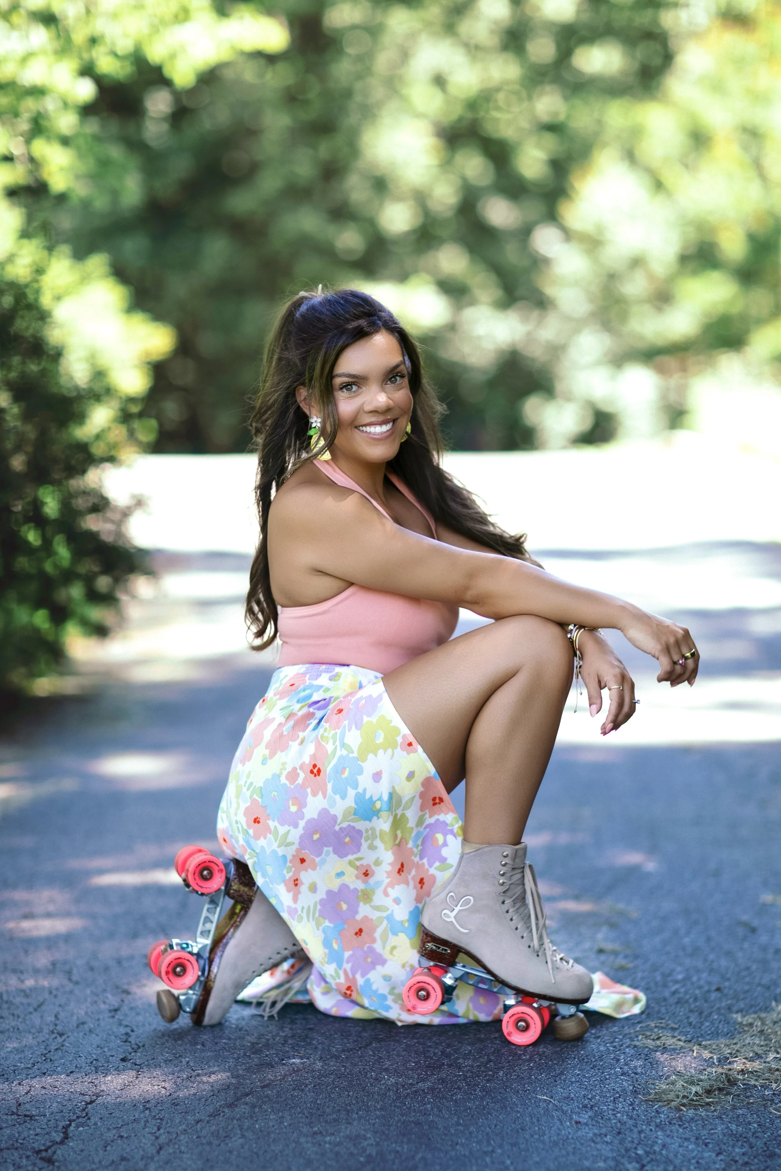 A young woman with long dark hair and bright eyes, wearing a pink top, a floral skirt, and roller skates, sitting on the pavement outside surrounded by green trees, smiling at the camera.